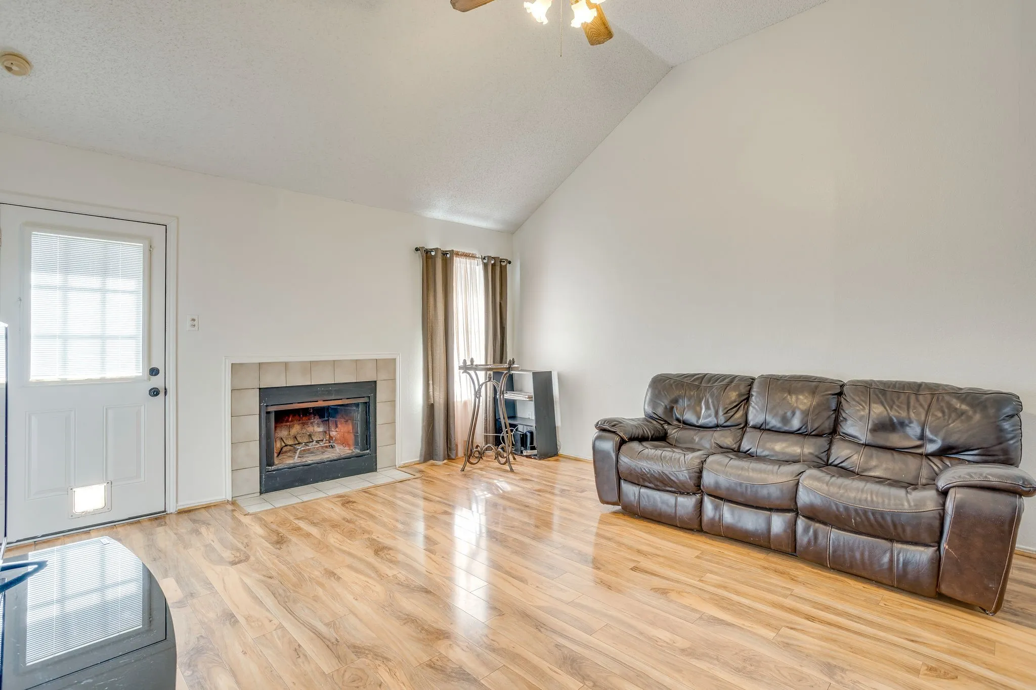 Vaulted ceiling, wood-finish flooring, fireplace with tile surround, white solid-panel door with window, ceiling fan with light fixture