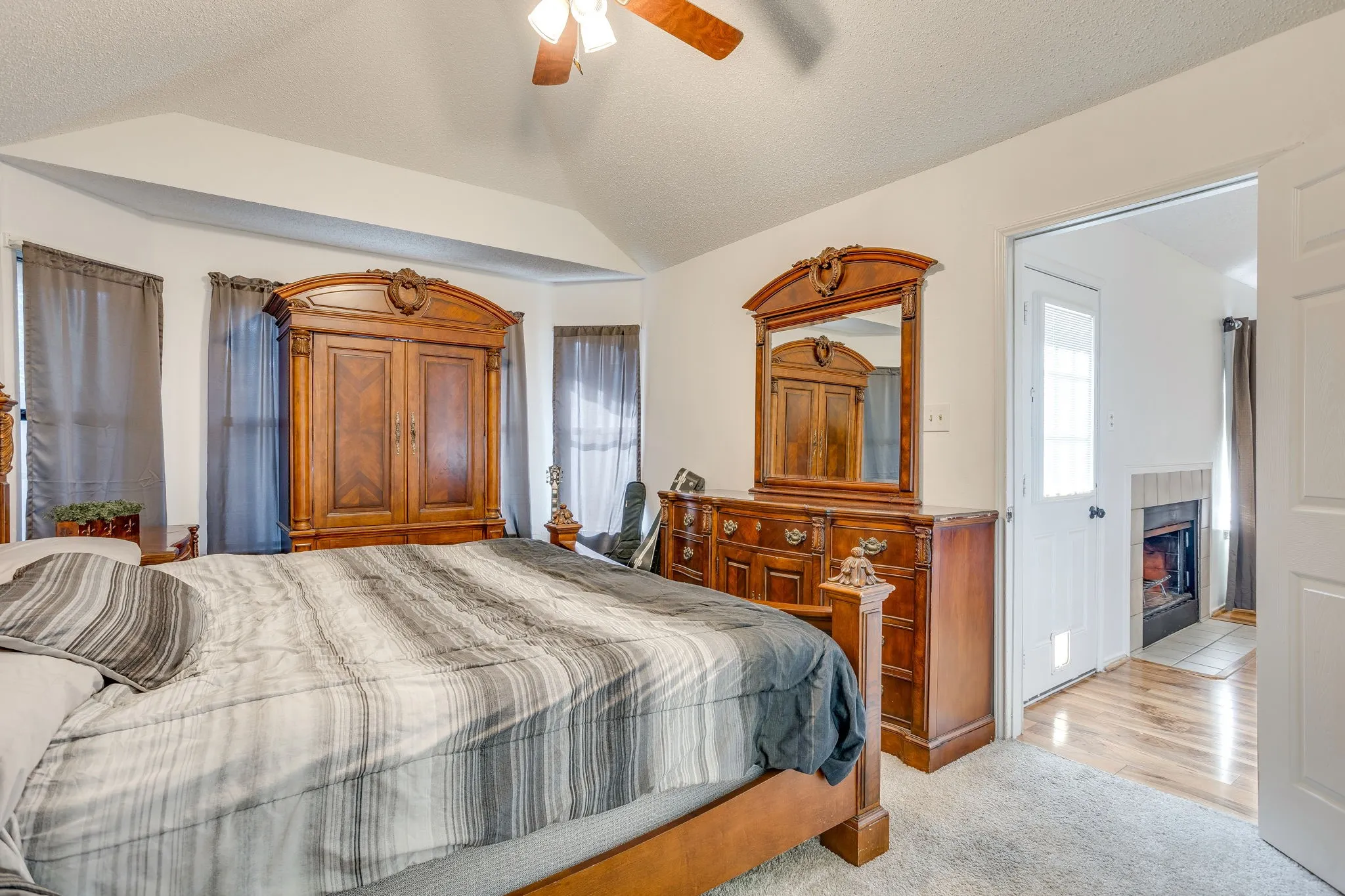 Vaulted ceiling with ceiling fan, bay window with multiple windows, carpet flooring, and an adjoining space with wood-finish flooring and a fireplace