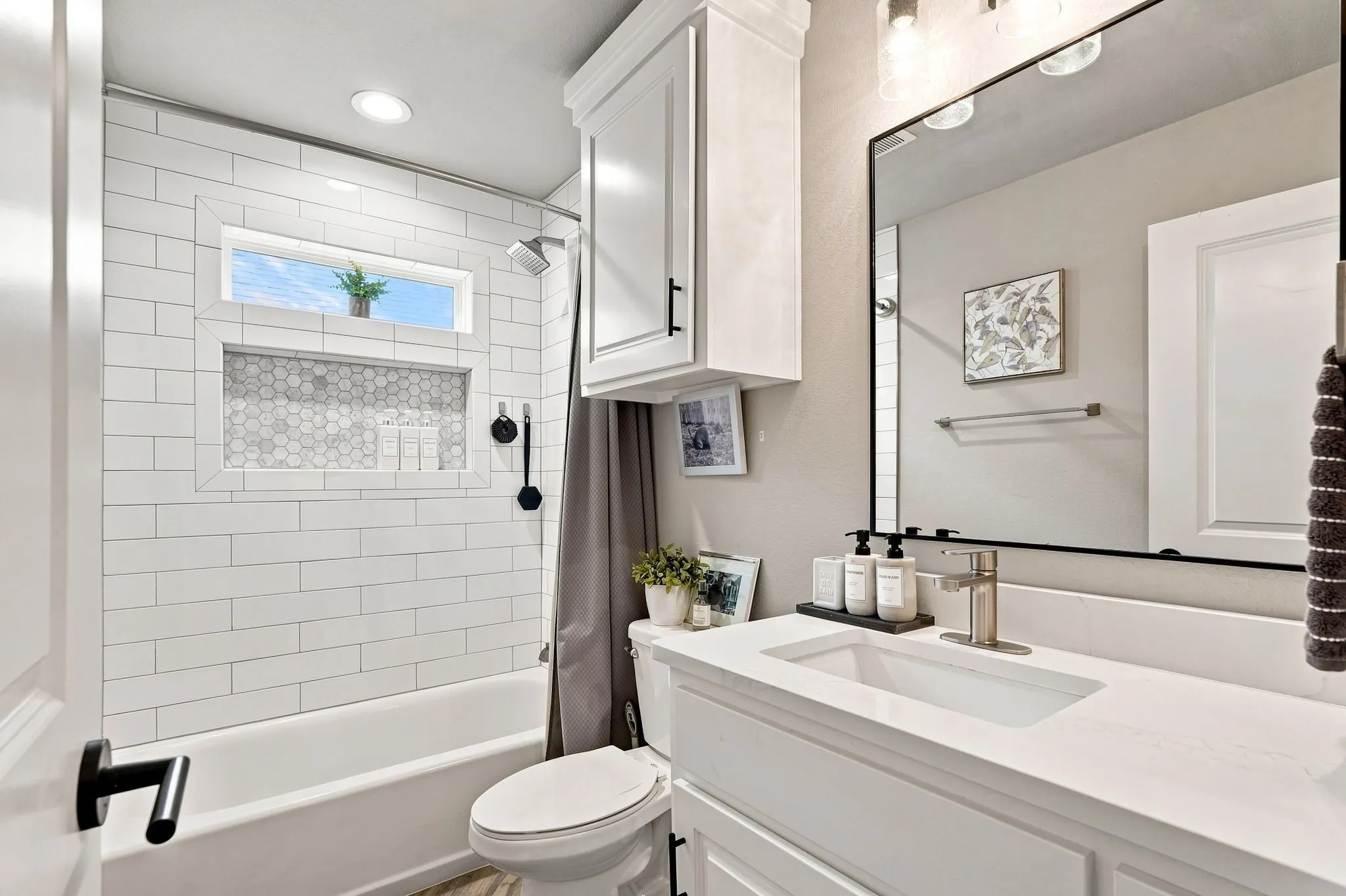 White subway tile shower surround with hexagon tile niche and transom window, white vanity with integrated sink and chrome faucet, large rectangular mirror, and white built-in cabinetry