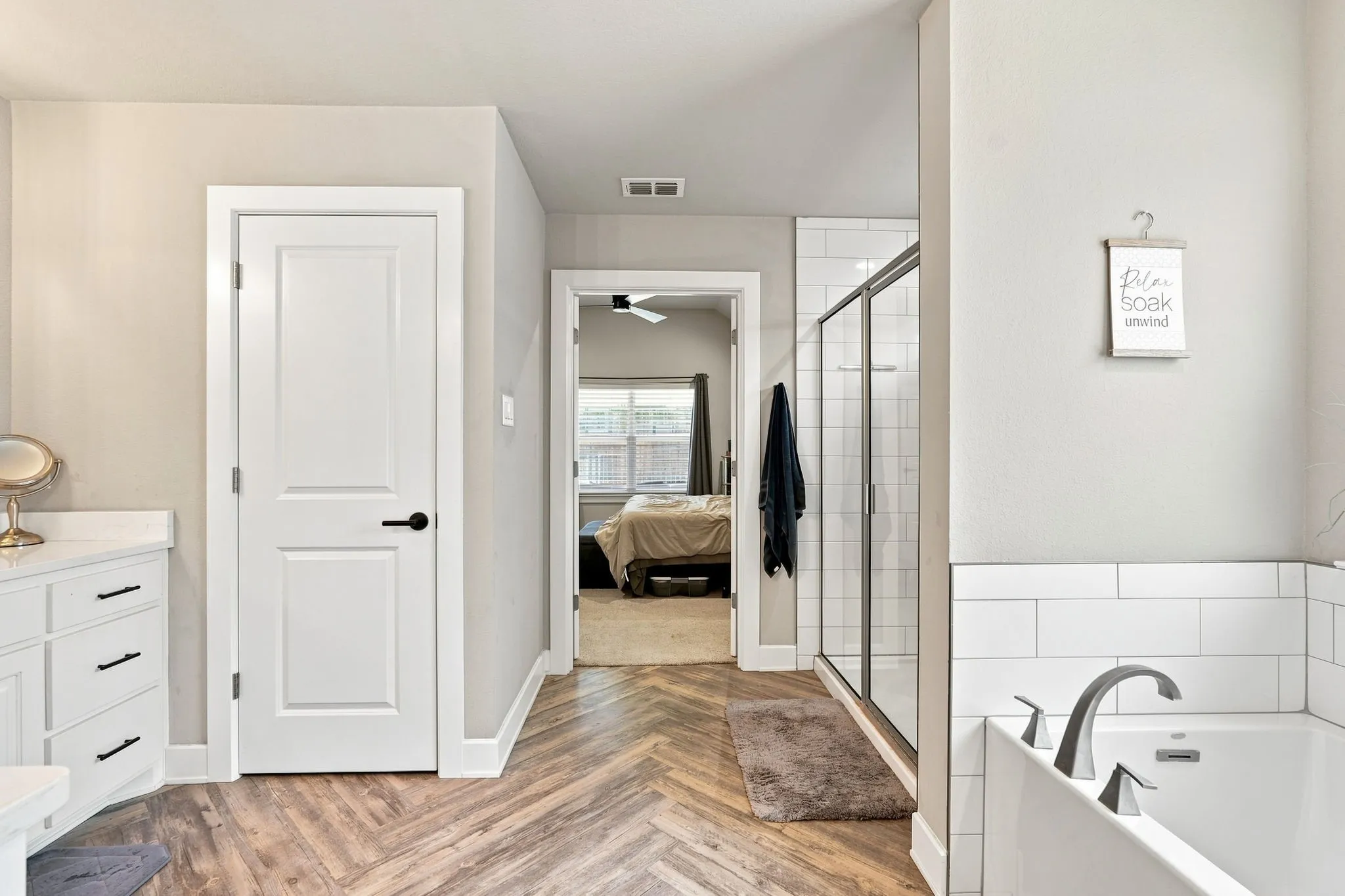 Wood-finish flooring in a herringbone pattern, white subway tile accents, a walk-in shower with clear glass enclosure, a built-in bathtub with contemporary fixtures, and a vanity with a white countertop and dark hardware