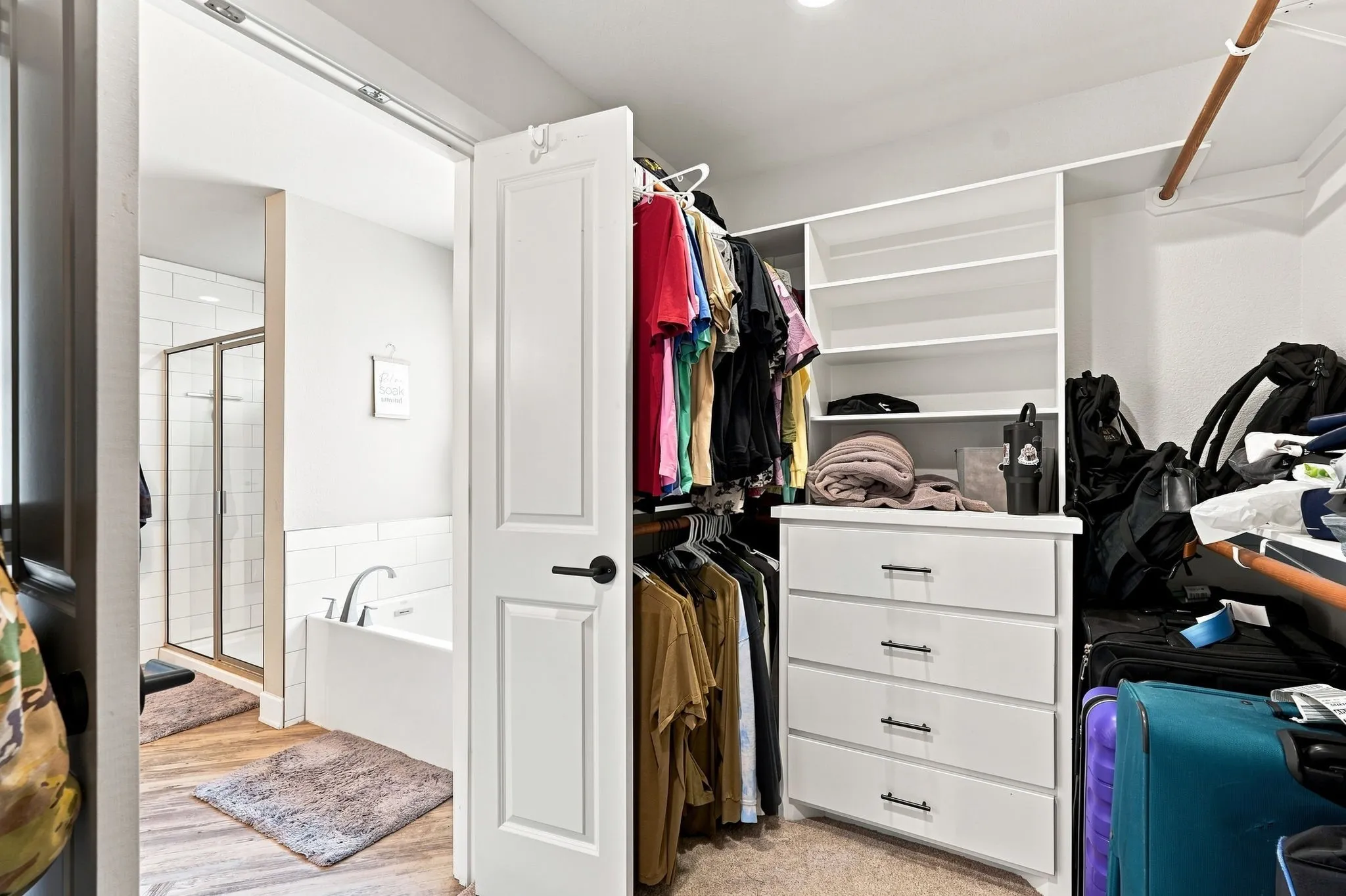 Built-in closet shelving, integrated dresser with dark hardware, wood-finish flooring, white subway tile surround, and a glass-enclosed shower
