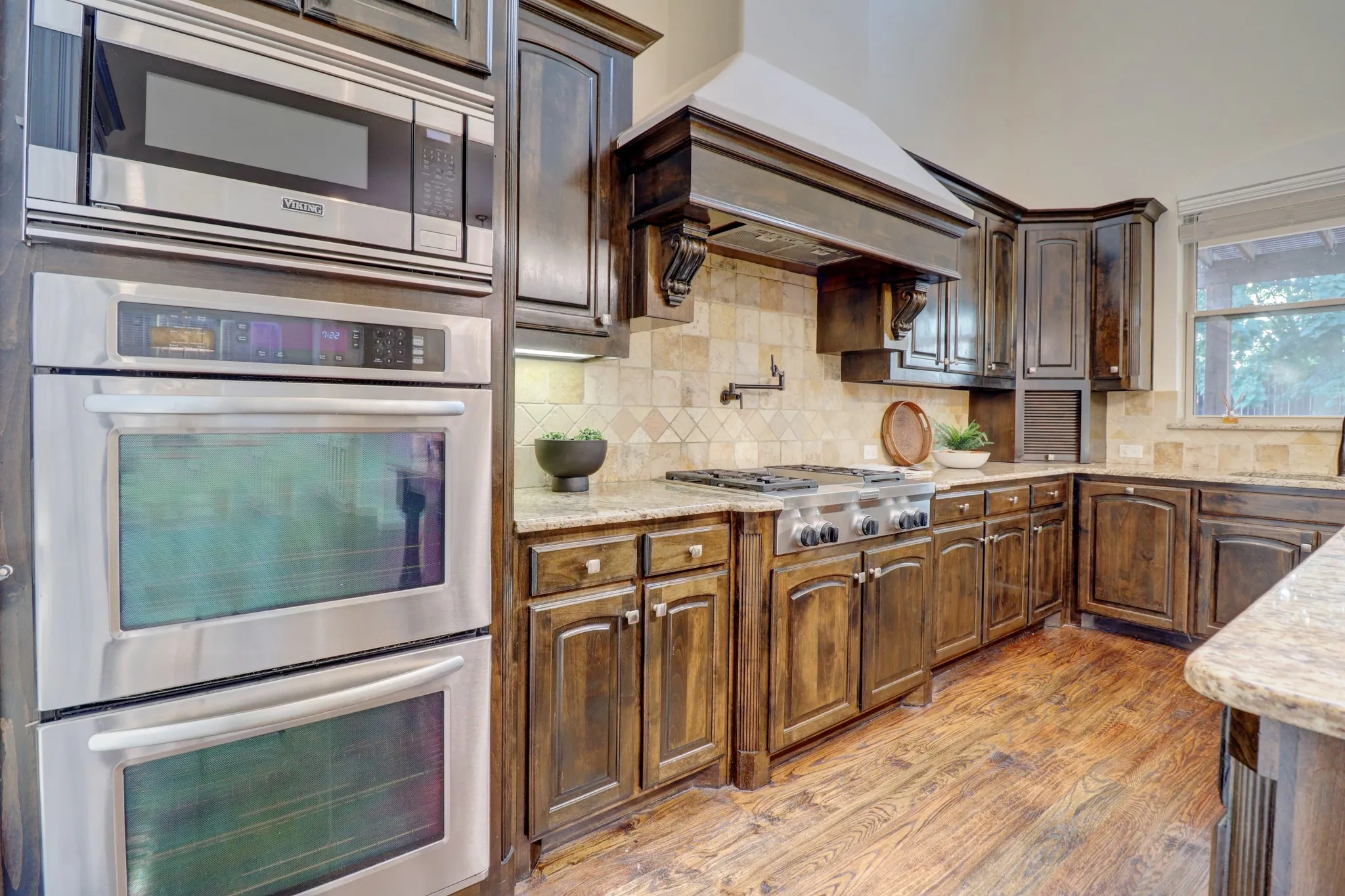 Dark wood-finish cabinetry, stainless steel double ovens, gas range with an ornate hood, tile backsplash, and light-toned countertops