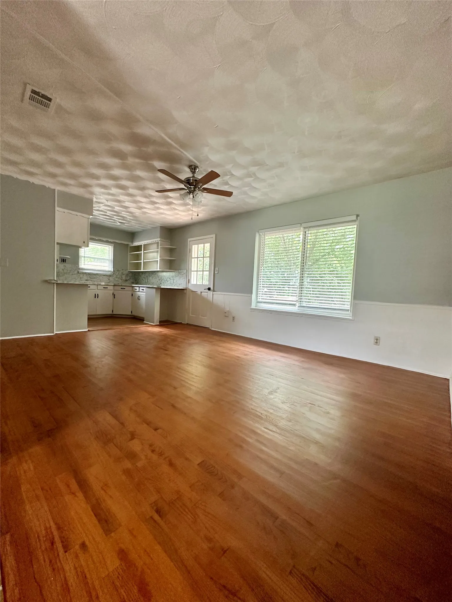 Hardwood flooring, ceiling fan, white wall cabinetry, white backsplash, and large window with blinds