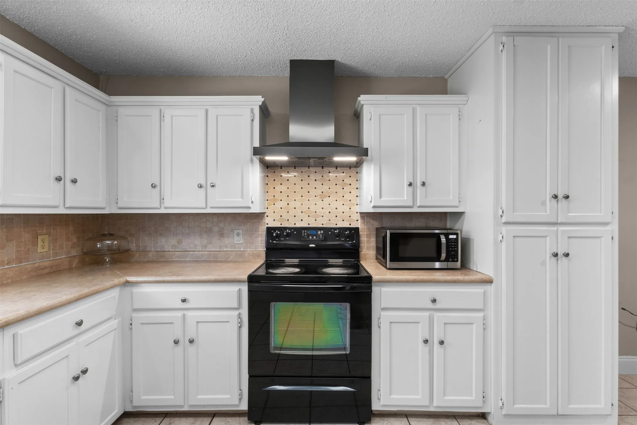 White cabinetry with crown molding, stainless steel range hood, built-in pantry storage, speckled backsplash, and light-toned countertops