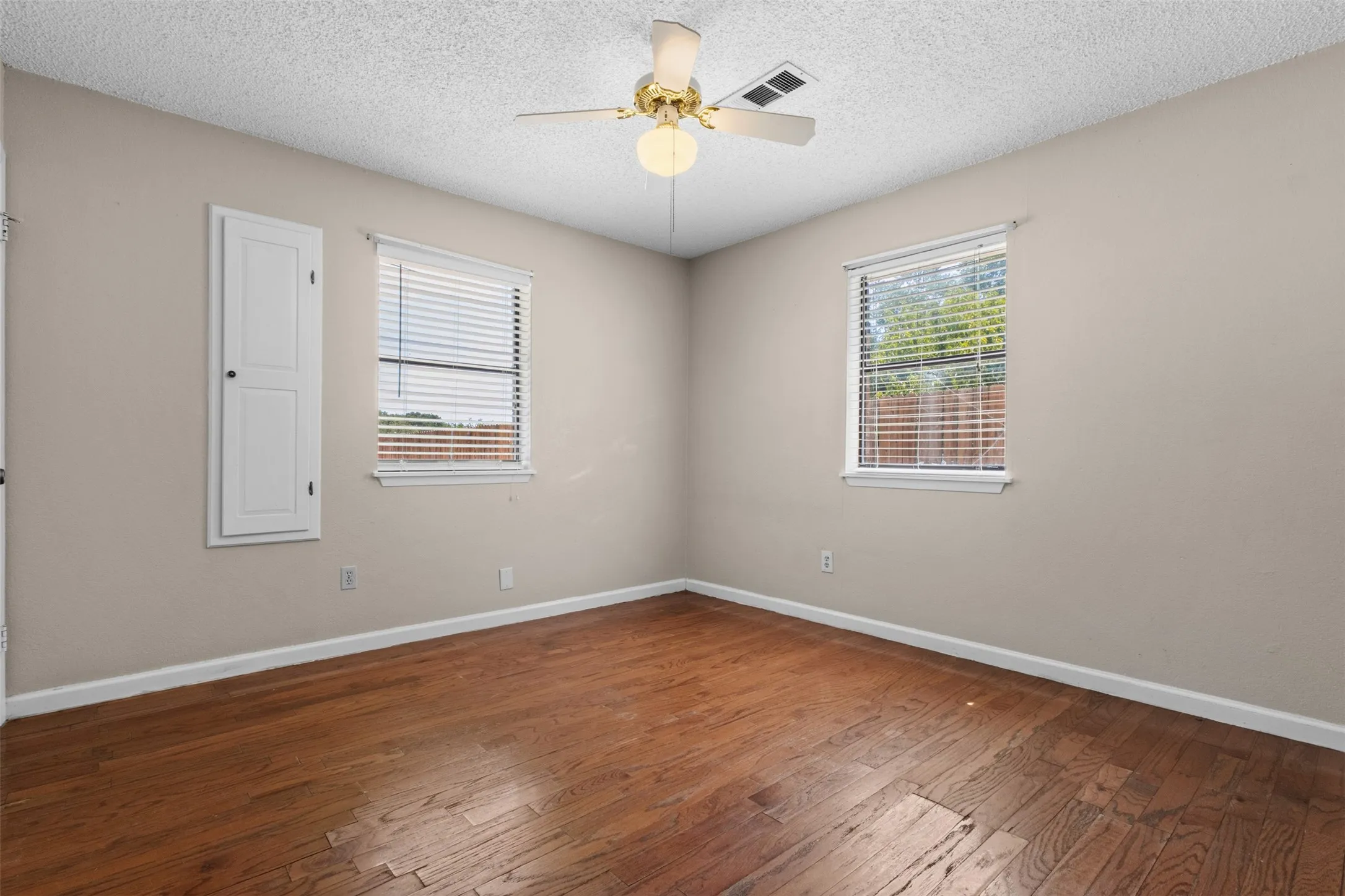 Rich wood-finish flooring, two windows with blinds, a ceiling fan with integrated lighting, and a built-in cabinet