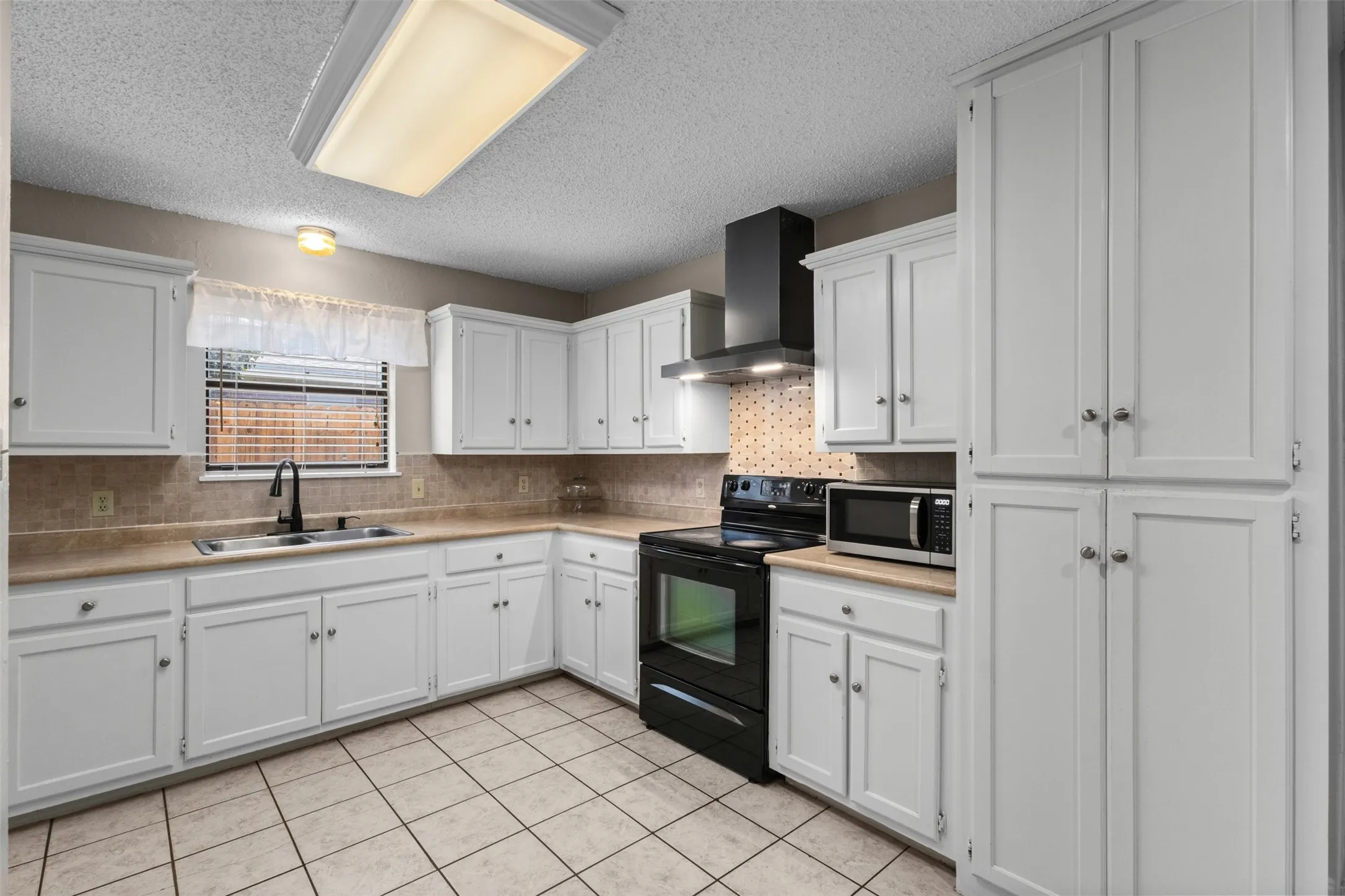 White paneled cabinetry, L-shaped countertop, black range with range hood, stainless steel sink with matte black faucet, and tiled flooring
