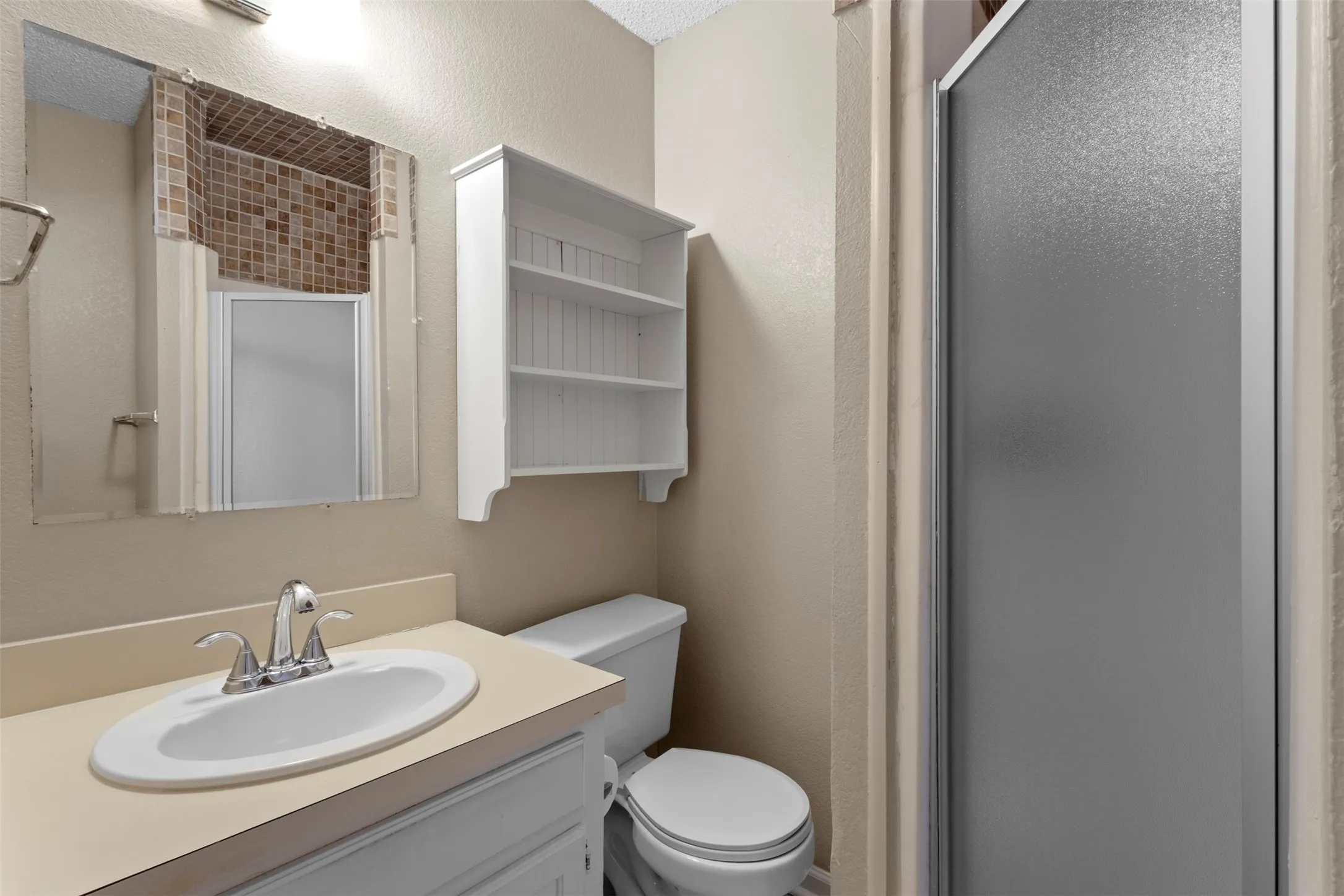 Bathroom featuring a vanity with an integrated sink, chrome faucet, wall-mounted mirror, and an adjacent shower with a frosted glass door