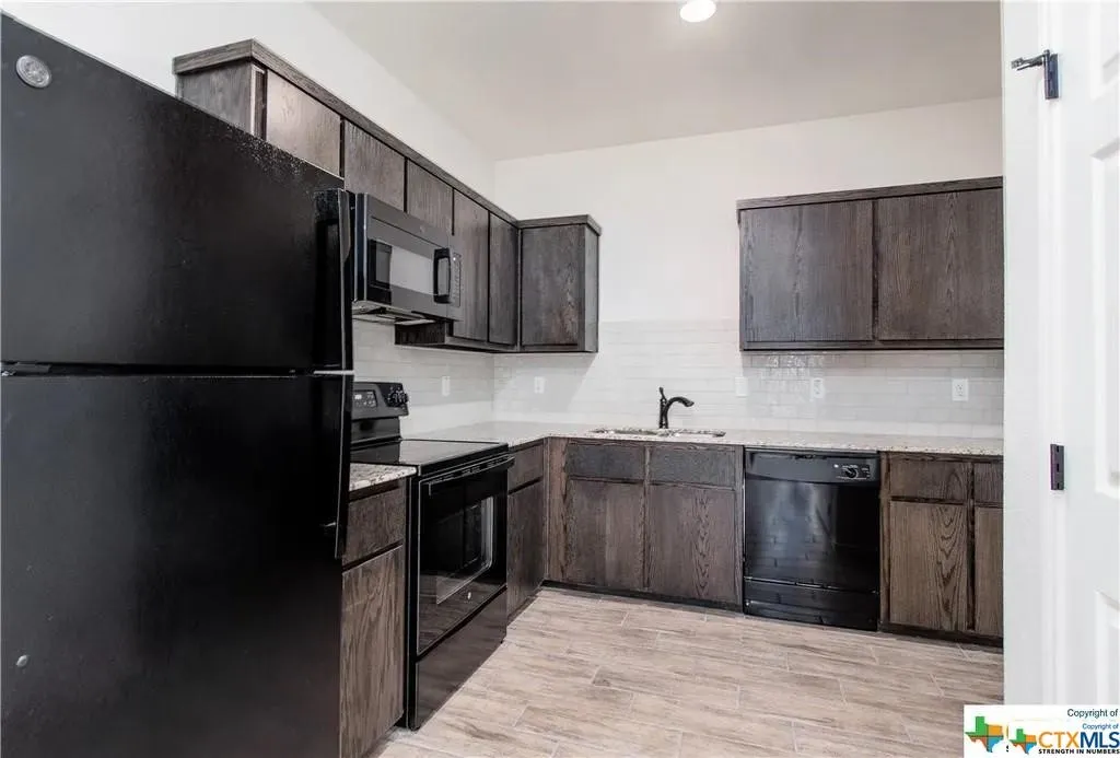 Contemporary kitchen featuring dark wood-finish cabinetry, black appliances, light-toned countertops, a white subway tile backsplash, and wood-look tile flooring