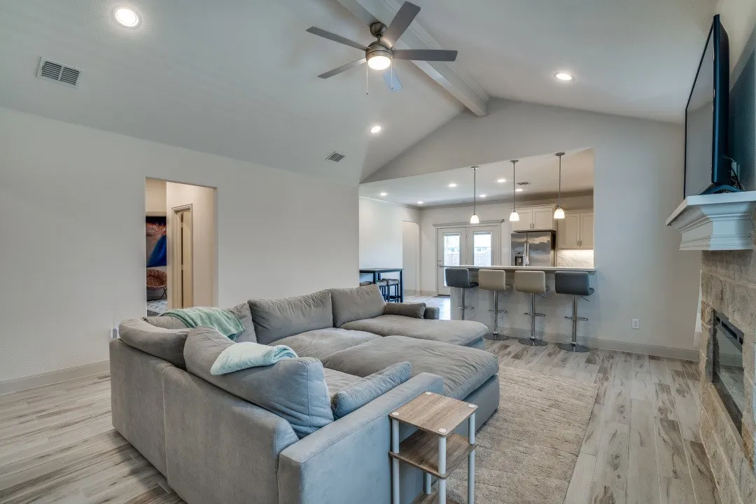 Vaulted ceiling with exposed beam, recessed lighting, wood-finish flooring, stone-clad fireplace with mantel, and a kitchen pass-through with pendant lighting