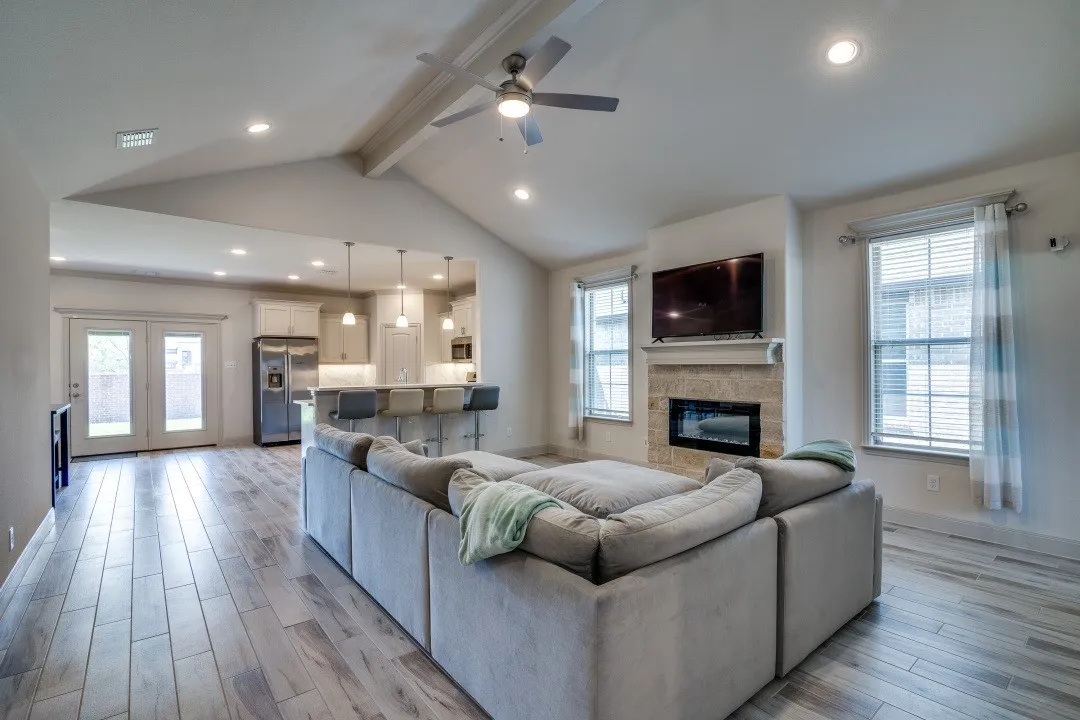 Vaulted ceiling with exposed beam, recessed lighting, stone-clad fireplace, wood-finish flooring, and French doors