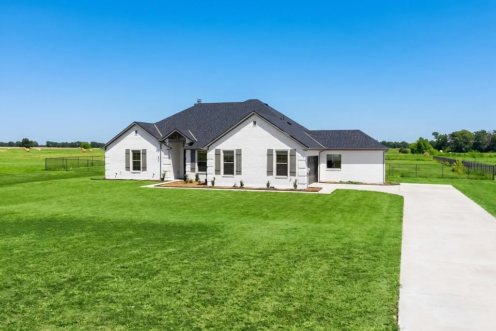 White brick exterior with dark shutters, dark shingle roof, expansive green lawn, and a concrete driveway