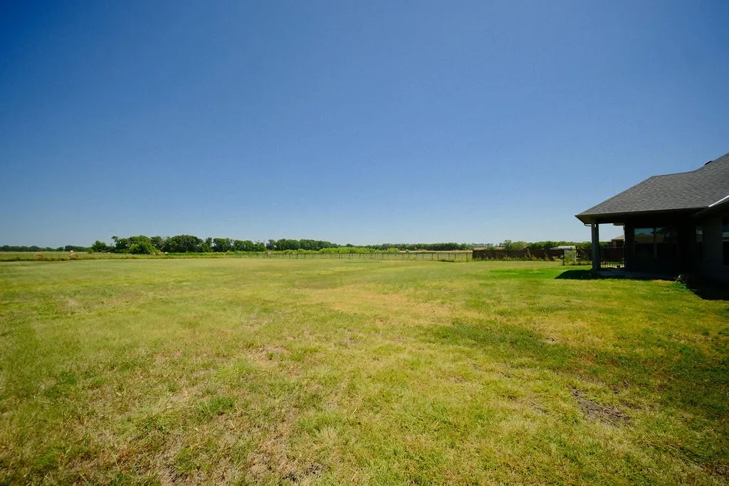Expansive grass lawn with a covered patio, dark roof, and distant tree line