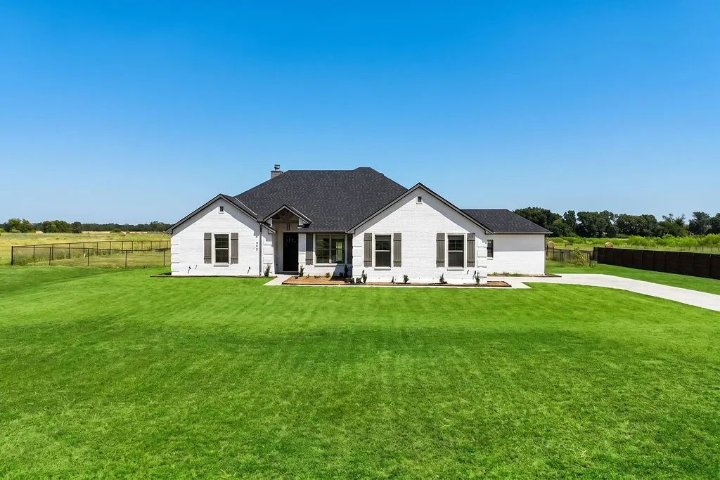 White brick exterior with dark shutters, a dark shingle roof, a concrete driveway, and a spacious green lawn