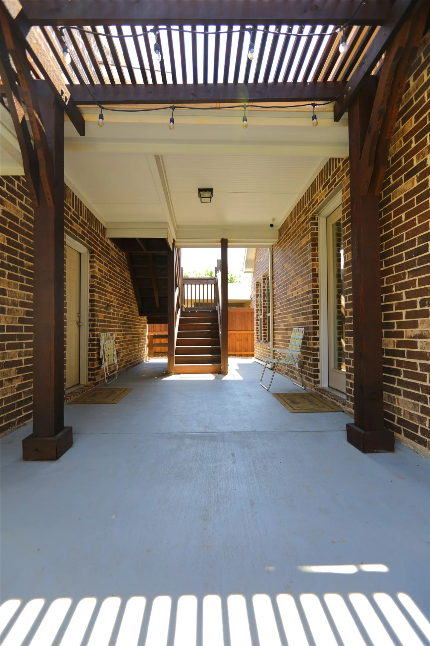 Spacious patio area featuring a slatted wood pergola with string lighting, brick exterior walls, and a light gray finished concrete surface