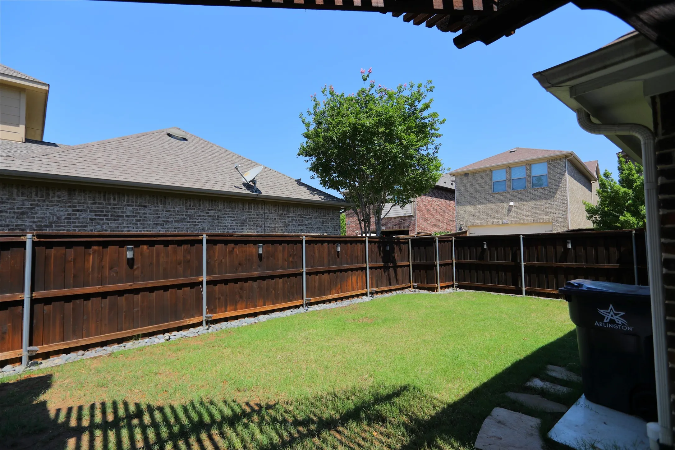 Grassed backyard with a privacy fence featuring integrated lighting, stone paver accents, and a covered patio area