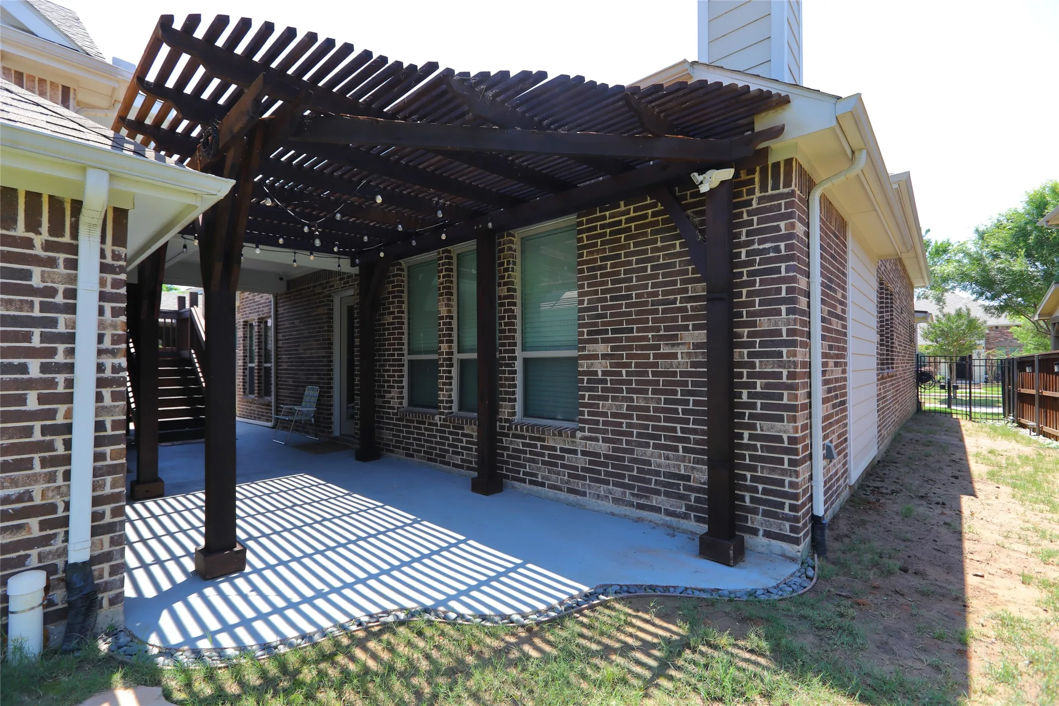 Expansive covered patio featuring a dark wood pergola, string lighting, brick exterior, concrete flooring, and a partial black metal fence