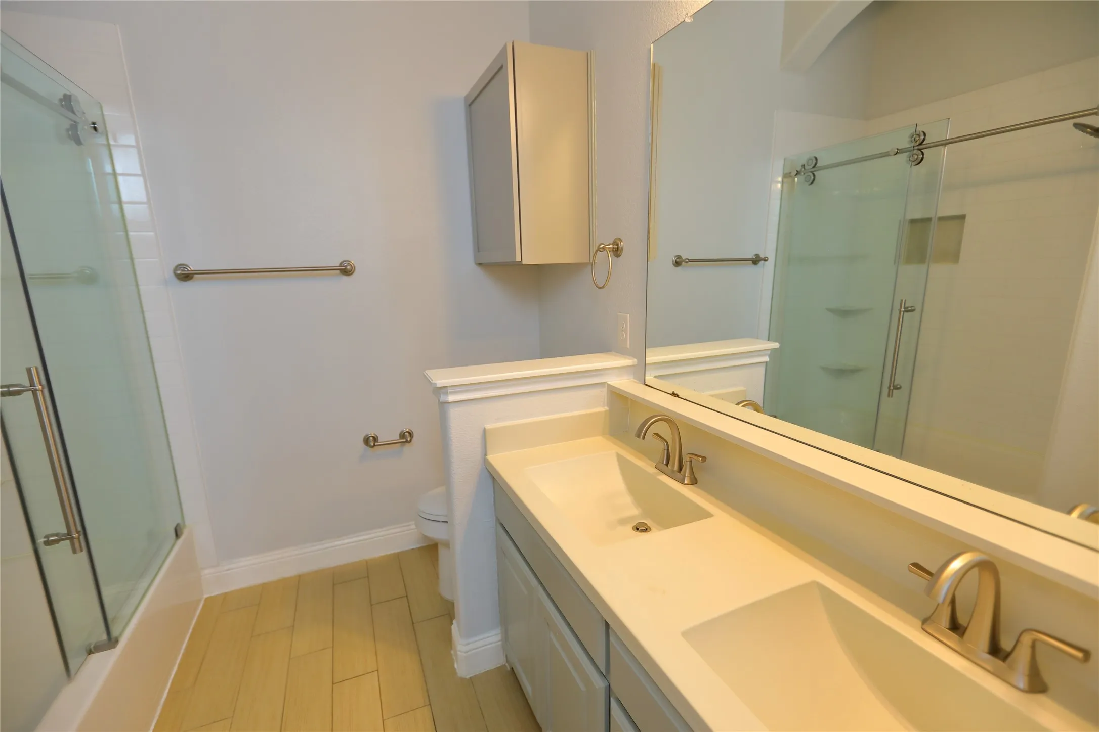 Bathroom featuring a dual vanity with integrated sinks and brushed nickel faucets, a full-width mirror, light wood-finish flooring, and a glass-enclosed shower/tub combination