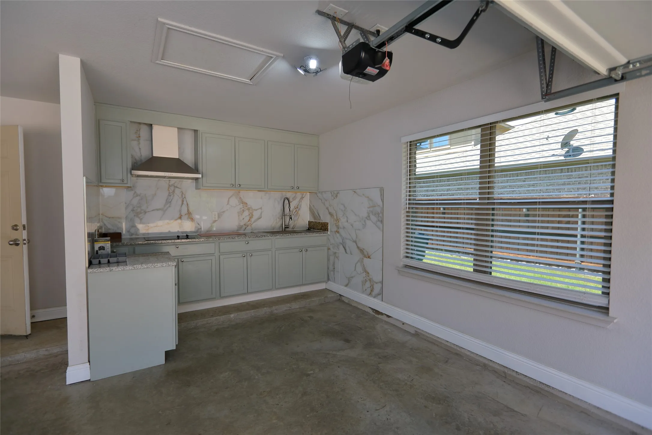 Kitchenette featuring light gray cabinetry, a stainless steel range hood, stone slab backsplash, and a window with horizontal blinds