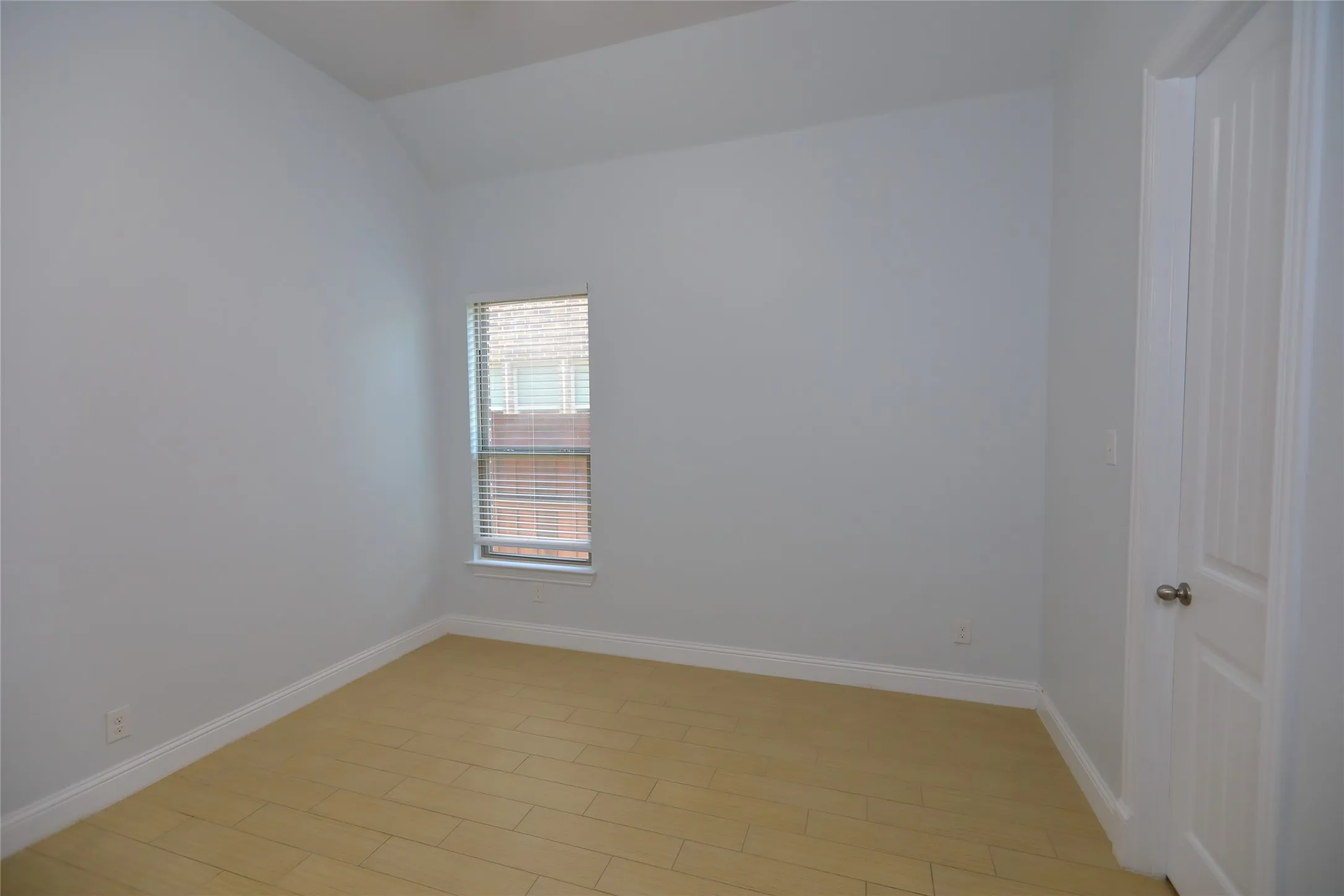 Room featuring light-toned wood-finish flooring, a single window with blinds, and a white paneled door with a brass-toned knob