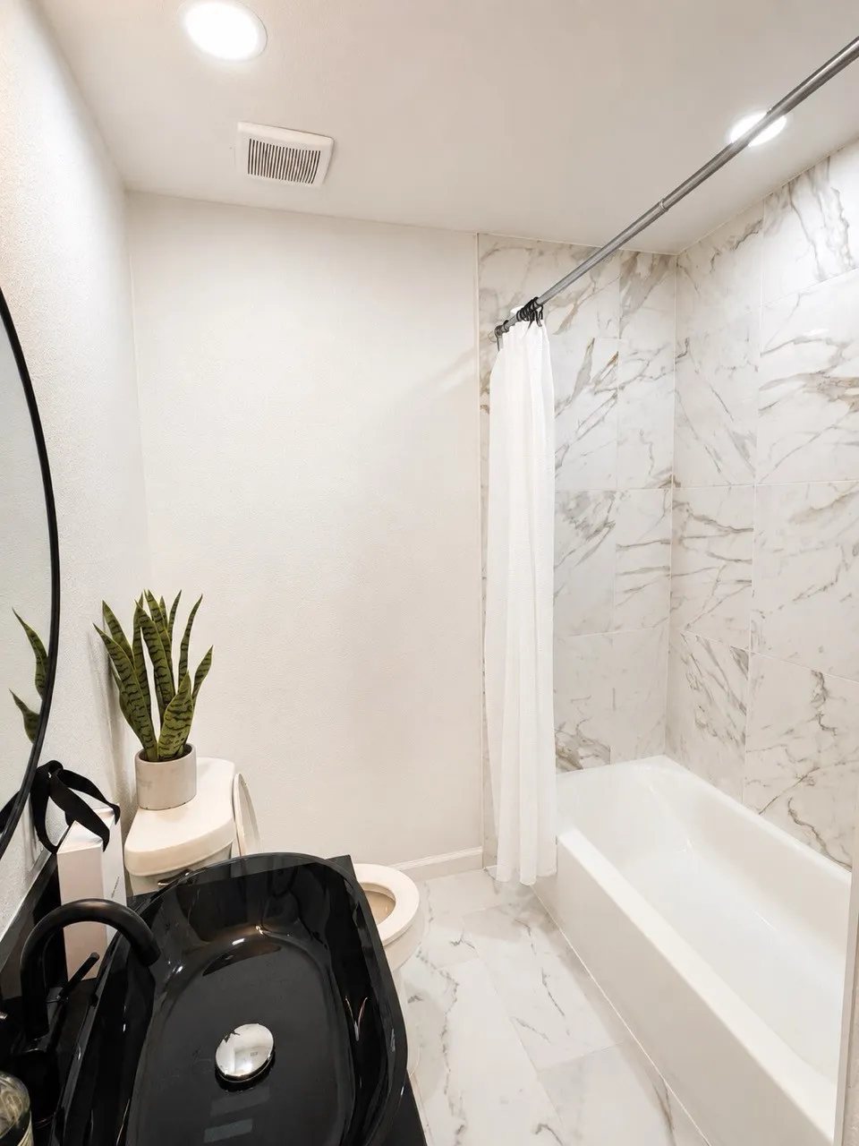 White bathtub with a white shower curtain, white and gray marble-patterned wall tile, white and gray marble-patterned floor tile, and a black vessel sink