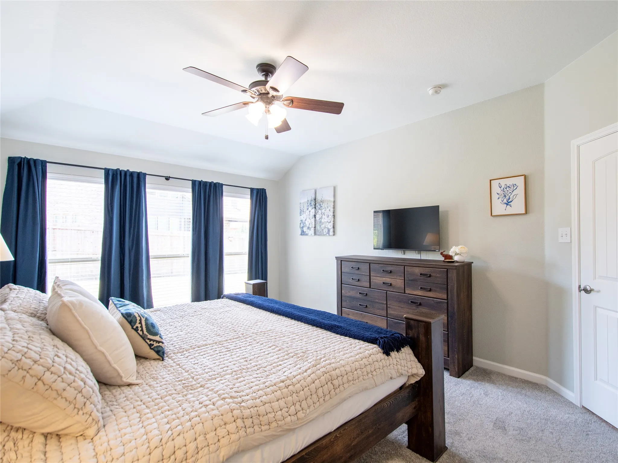 Vaulted ceiling with a ceiling fan, light gray wall paint, three large windows, carpet flooring, and a paneled door
