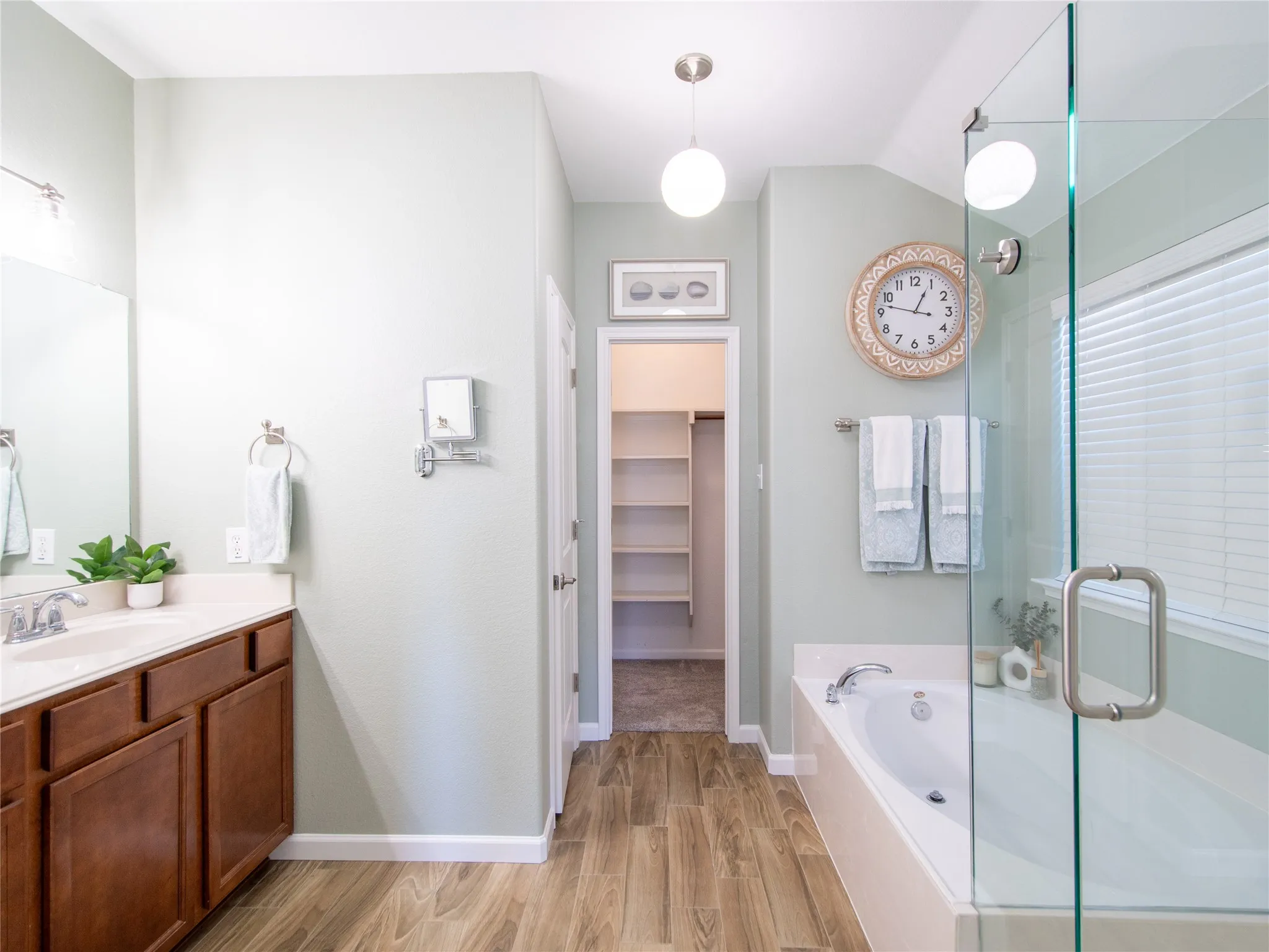 Bathroom featuring wood-finish flooring, a soaking tub, a glass-enclosed shower, a vanity with a white countertop and wood-finish cabinetry, and an open doorway leading to a closet with built-in shelving