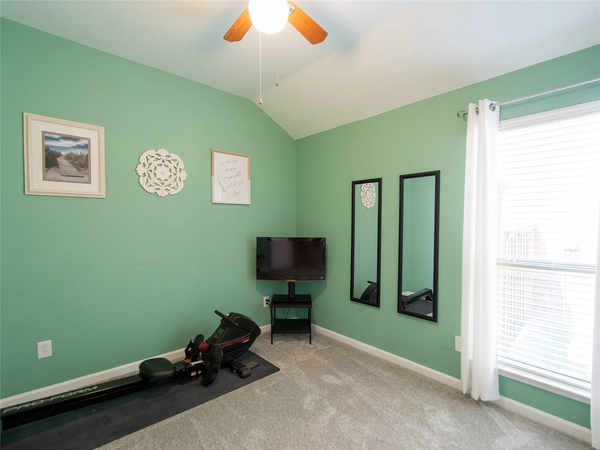 V-groove vaulted ceiling with ceiling fan, light-colored carpeting, large window with blinds and curtains, and green painted walls