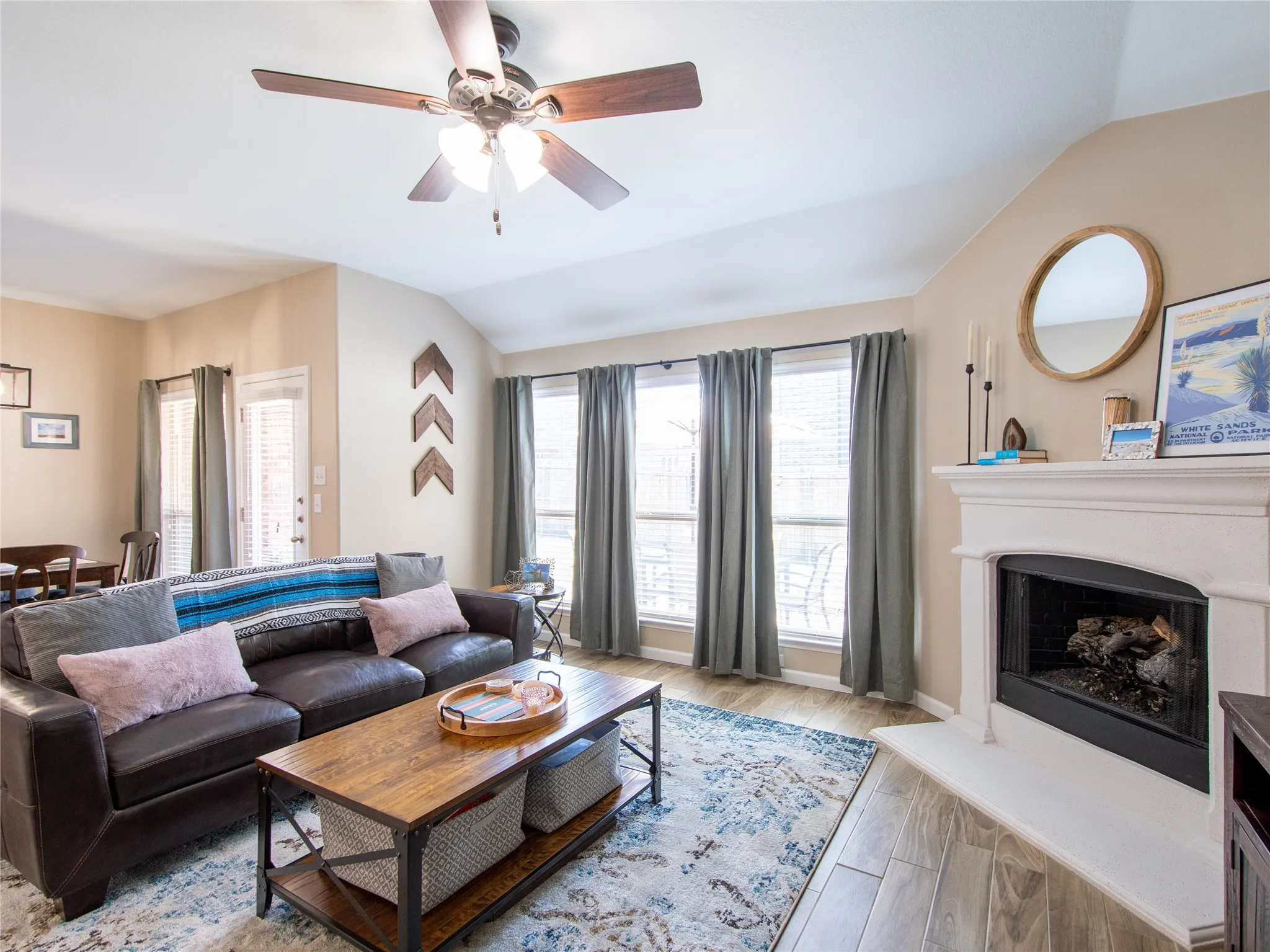 Vaulted ceilings with a ceiling fan, fireplace with a white mantel, wood-finish flooring, and multiple windows with vertical blinds