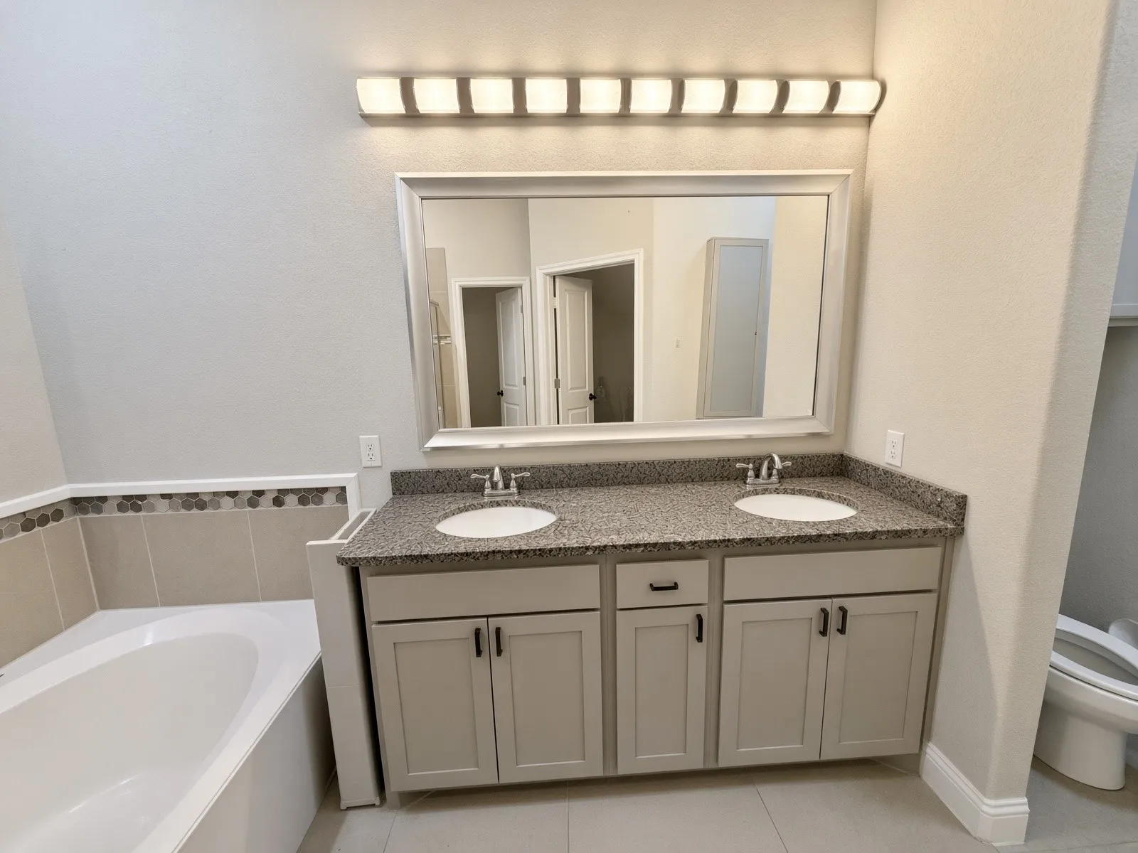 Dual vanity with granite-style countertop and integrated sinks, framed mirror, and a built-in bathtub with tiled surround