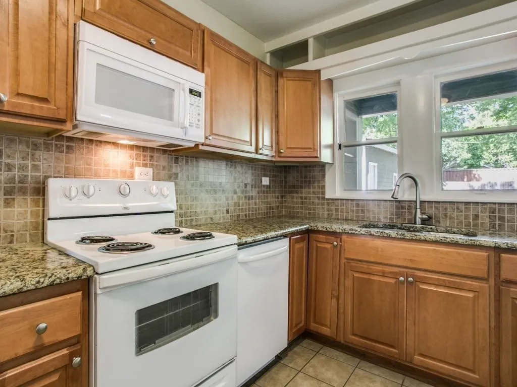 Wood cabinetry with white appliances, tile backsplash, stone countertops, and a dual-basin sink with a gooseneck faucet