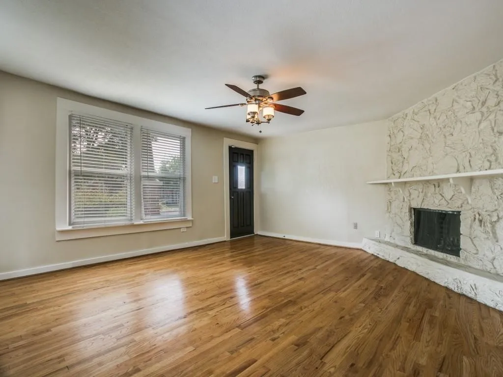 Hardwood flooring, stone fireplace, ceiling fan with integrated lighting, multiple windows with blinds, and a solid entry door with frosted glass