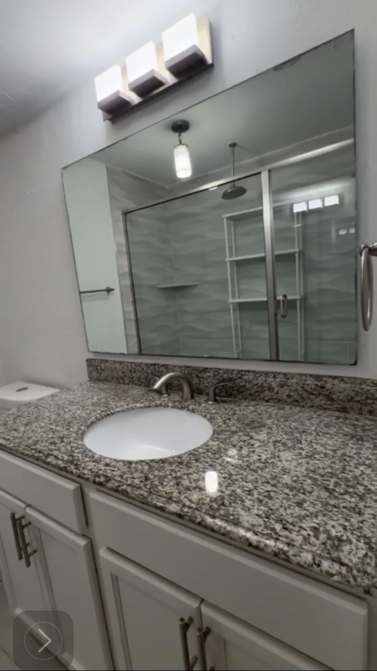 Spa-inspired bathroom featuring a large framed mirror, speckled stone countertop with an undermount circular sink, white cabinetry with brushed metal hardware, and contemporary lighting fixtures including a wall-mounted vanity light and pendant accent