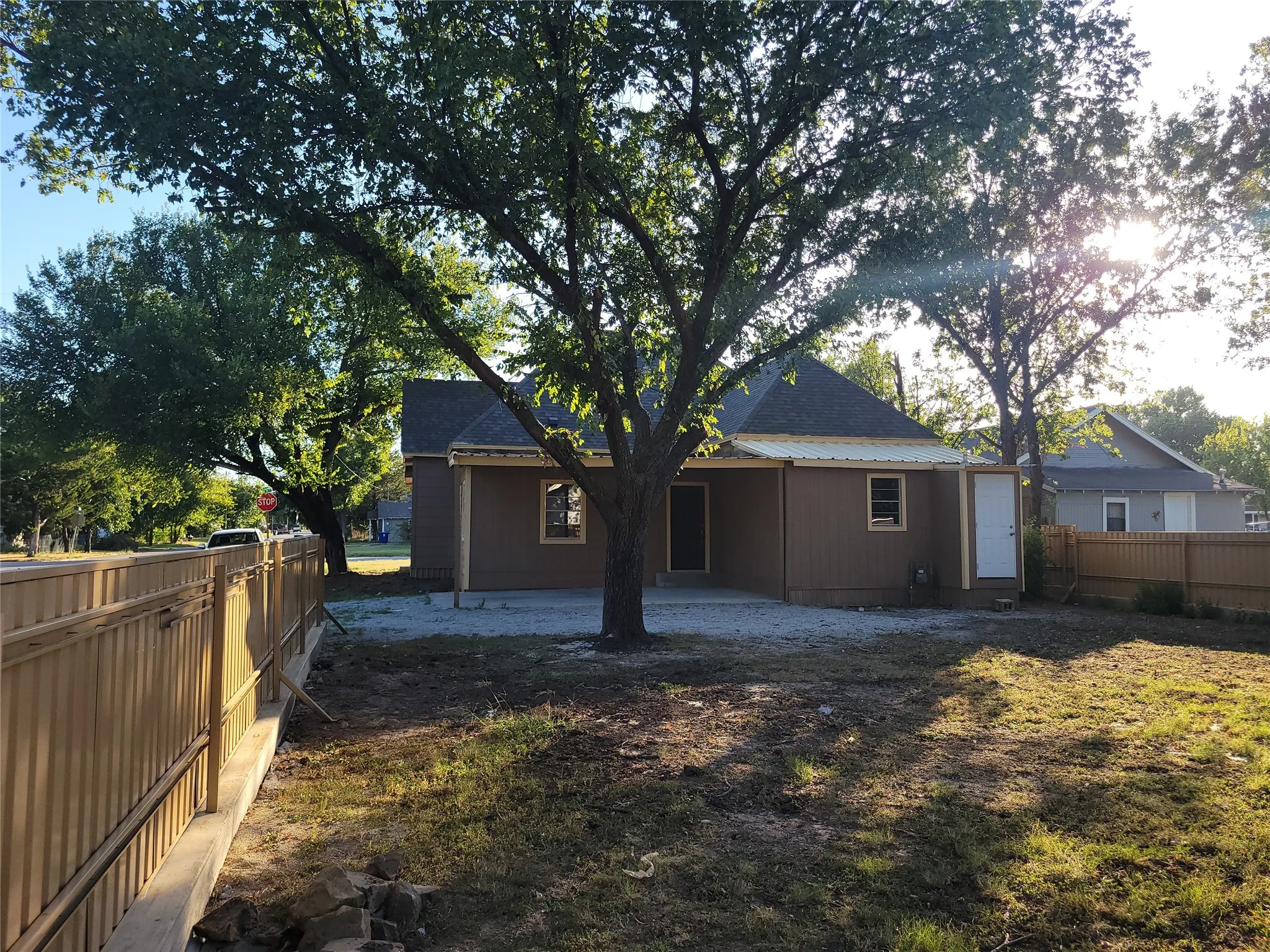 Shingle roof with brown siding, large mature trees, and a wooden privacy fence