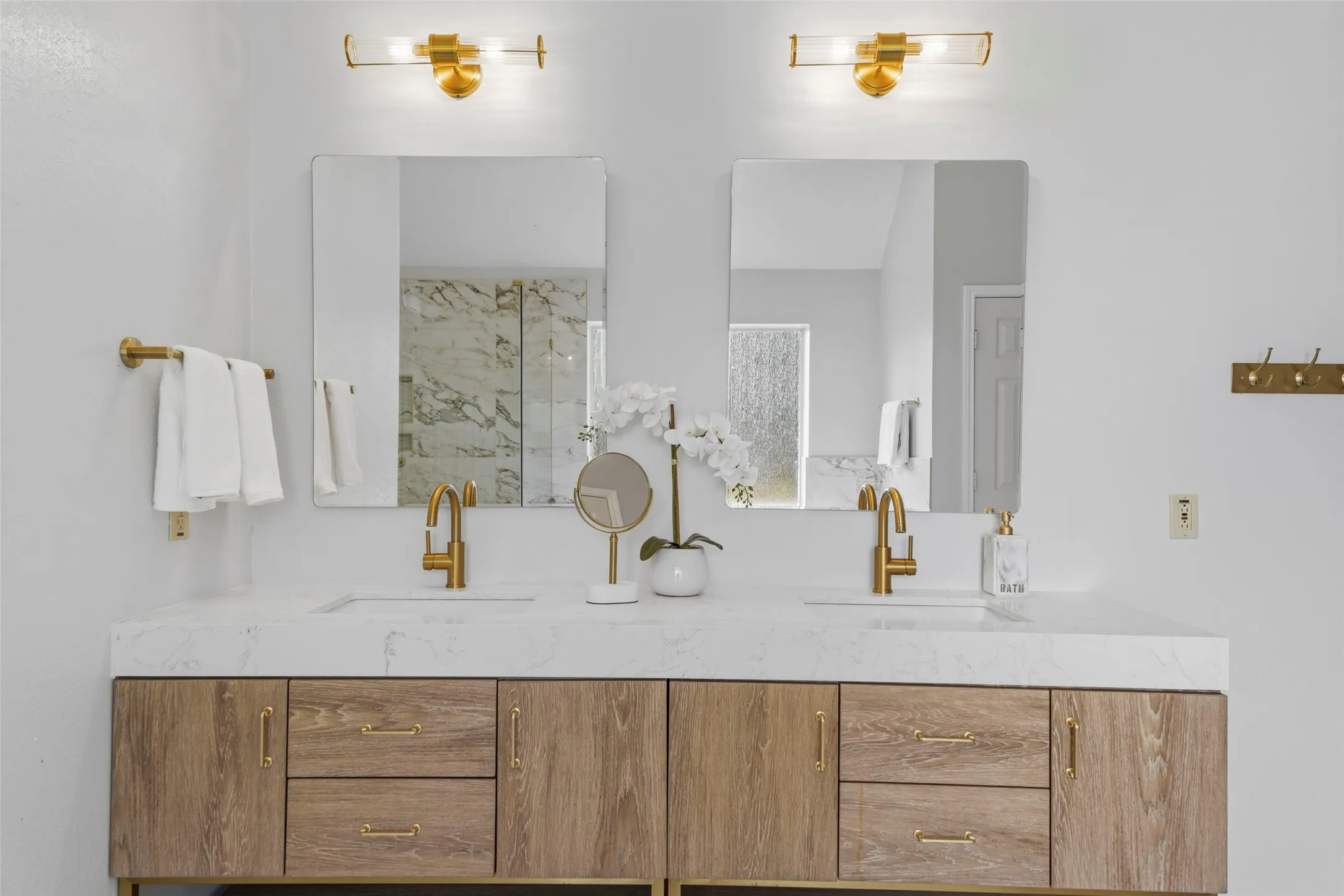 Dual vanity with integrated sinks, wood-finish cabinetry, sleek gold-tone hardware, and contemporary wall-mounted light fixtures