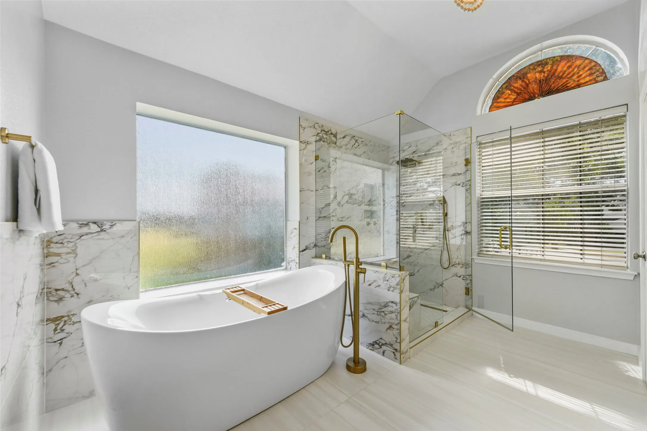 Freestanding soaking tub with a floor-mounted gold-toned faucet, glass-enclosed shower with marble-finish tile, and a decorative arched window