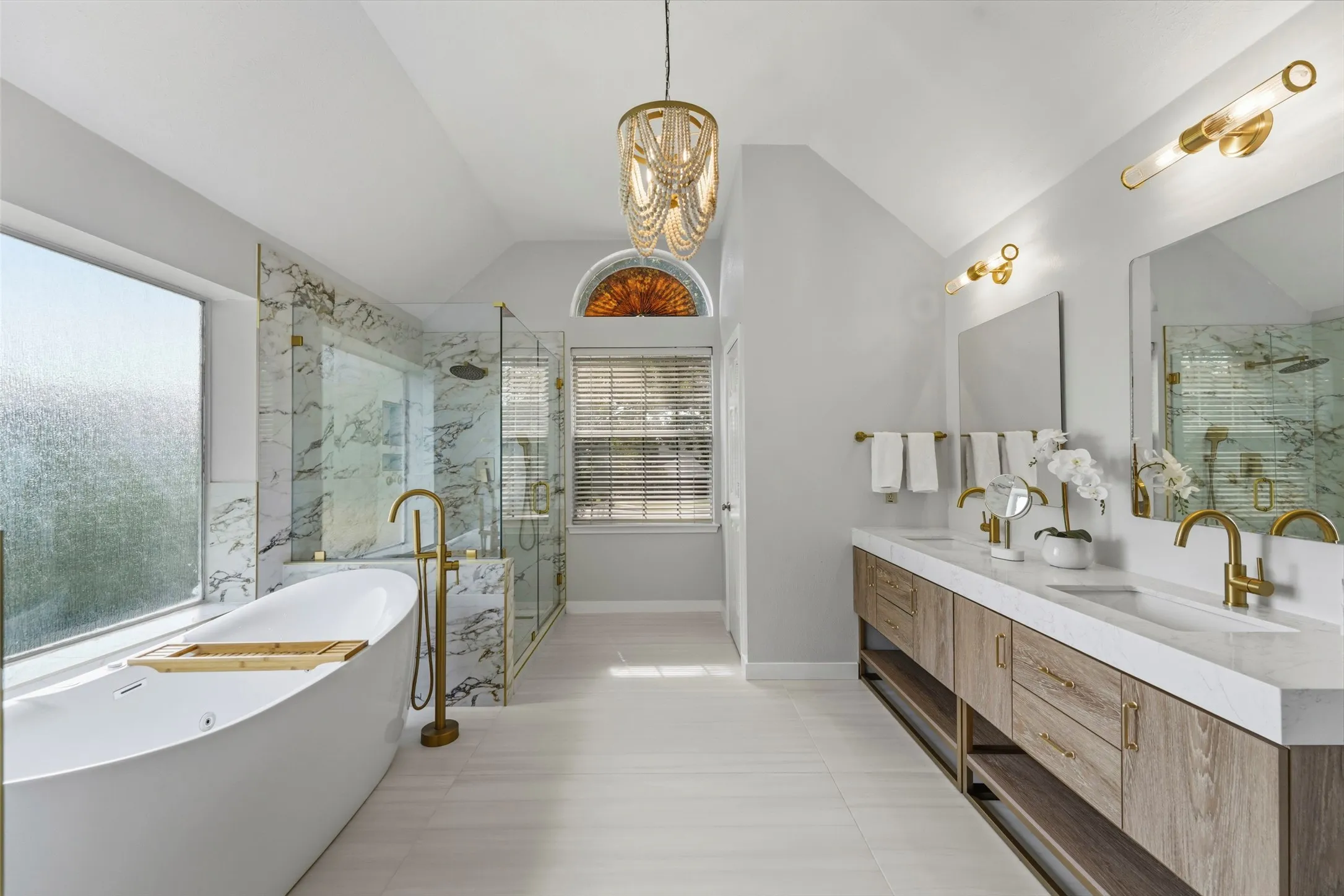 Vaulted ceiling, freestanding soaking tub with floor-mounted faucet, glass-enclosed shower with marble-finish tile, dual vanity with integrated sinks, and wood-finish cabinetry