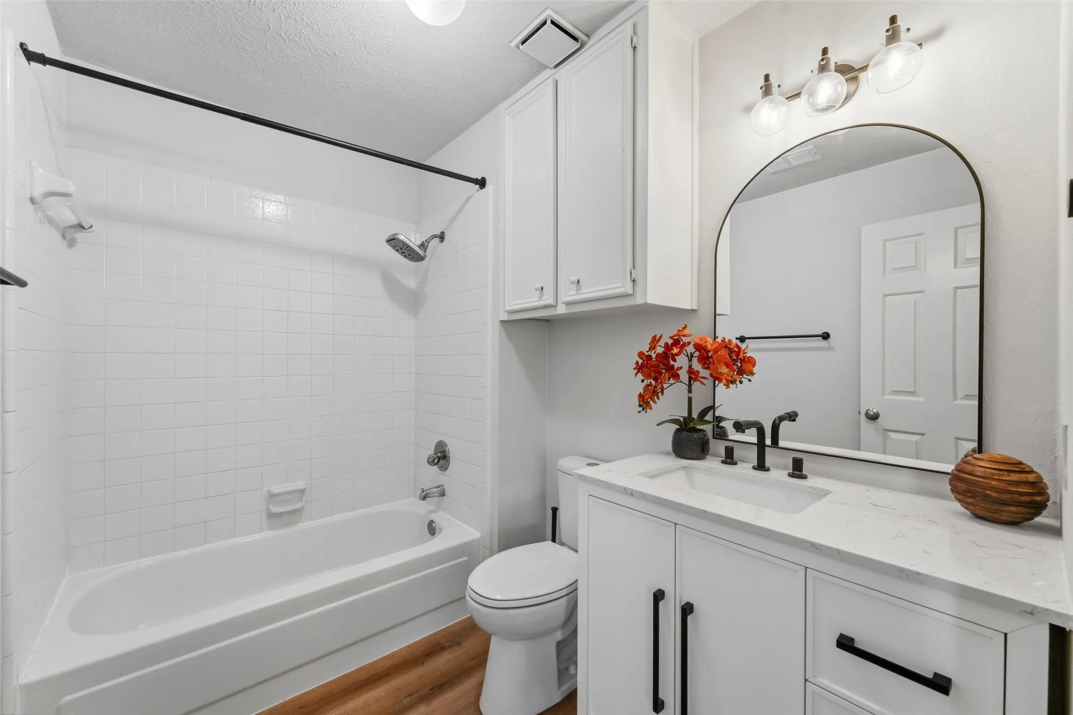 White tiled bath and shower combination, white vanity with integrated sink, arched mirror, matte black fixtures, and wood-finish flooring