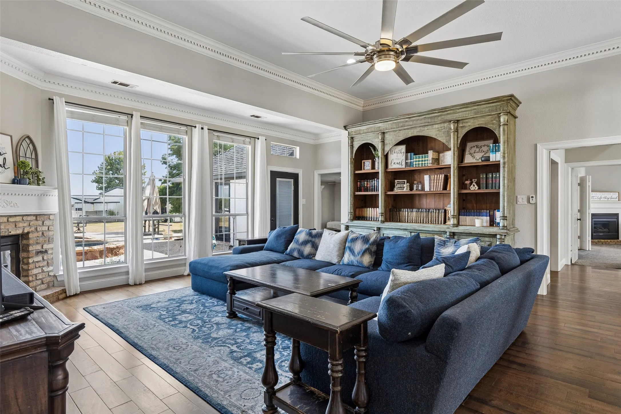 Expansive living room featuring large windows, a brick-surround fireplace, wood-finish flooring, a built-in bookshelf, and crown molding