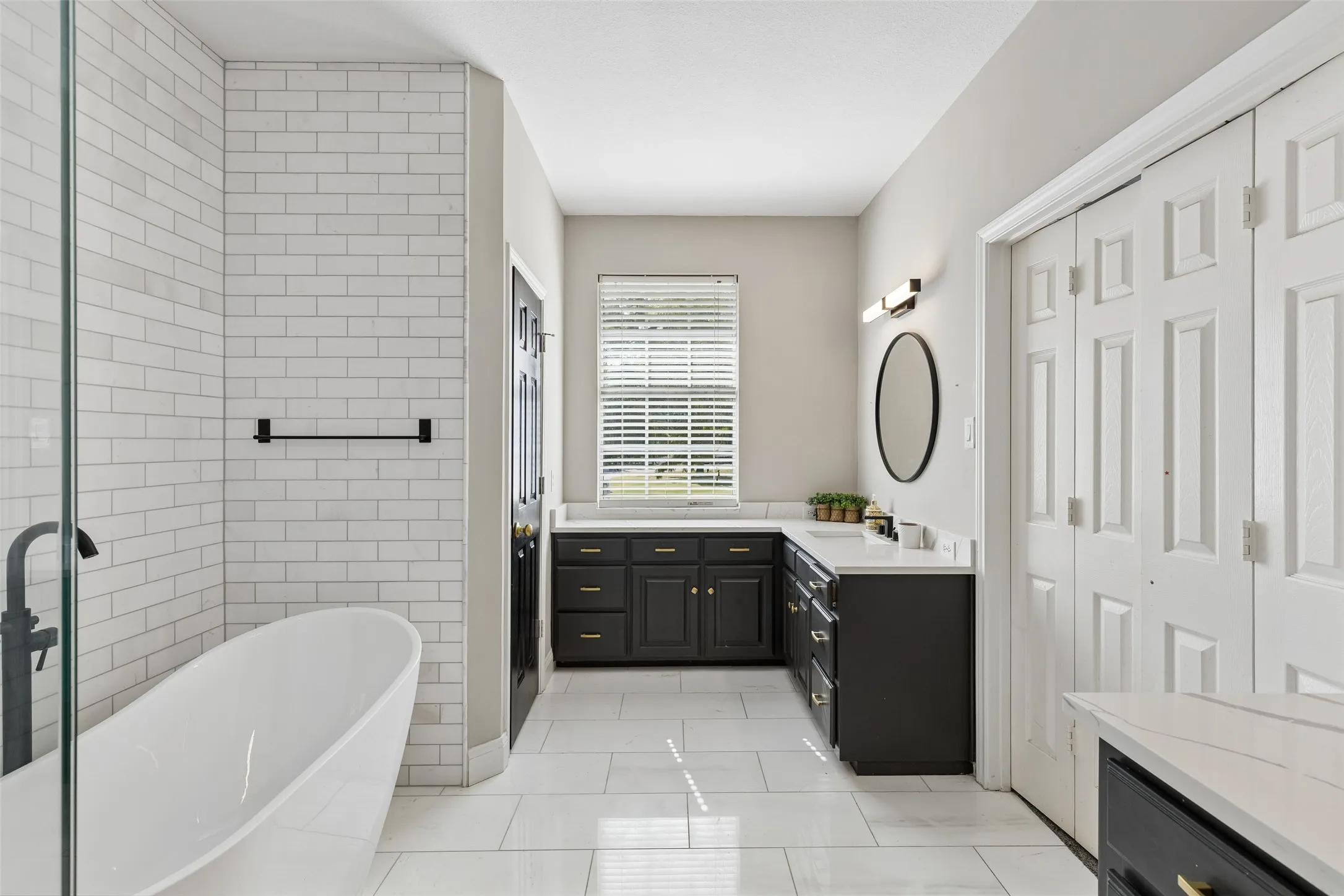 Freestanding bathtub with a black faucet, white subway tile shower surround, double vanity with dark cabinetry, and white quartz countertops