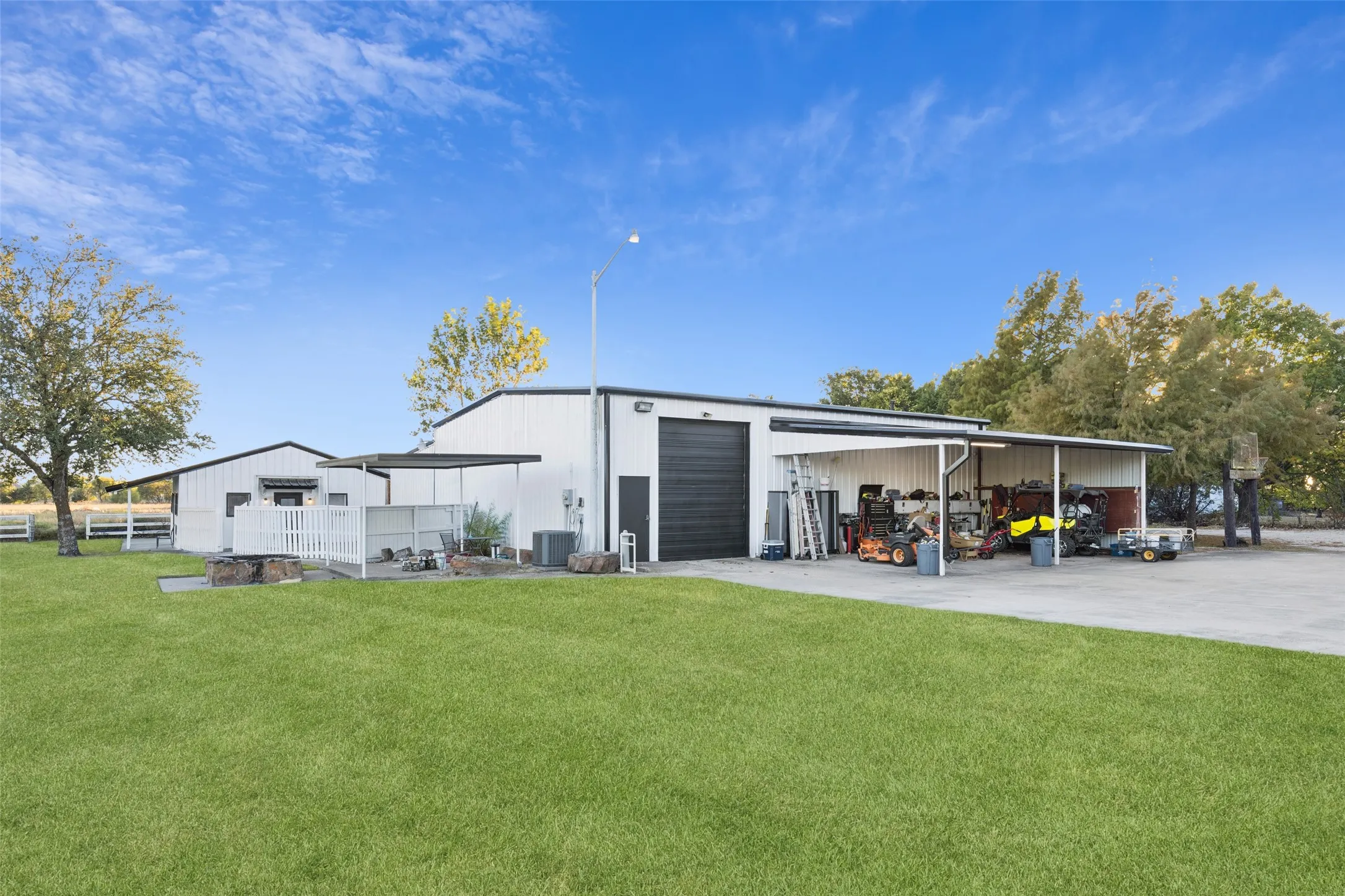 Expansive green space, white metal-sided buildings, large roll-up garage door, covered outdoor area, and paved concrete drive