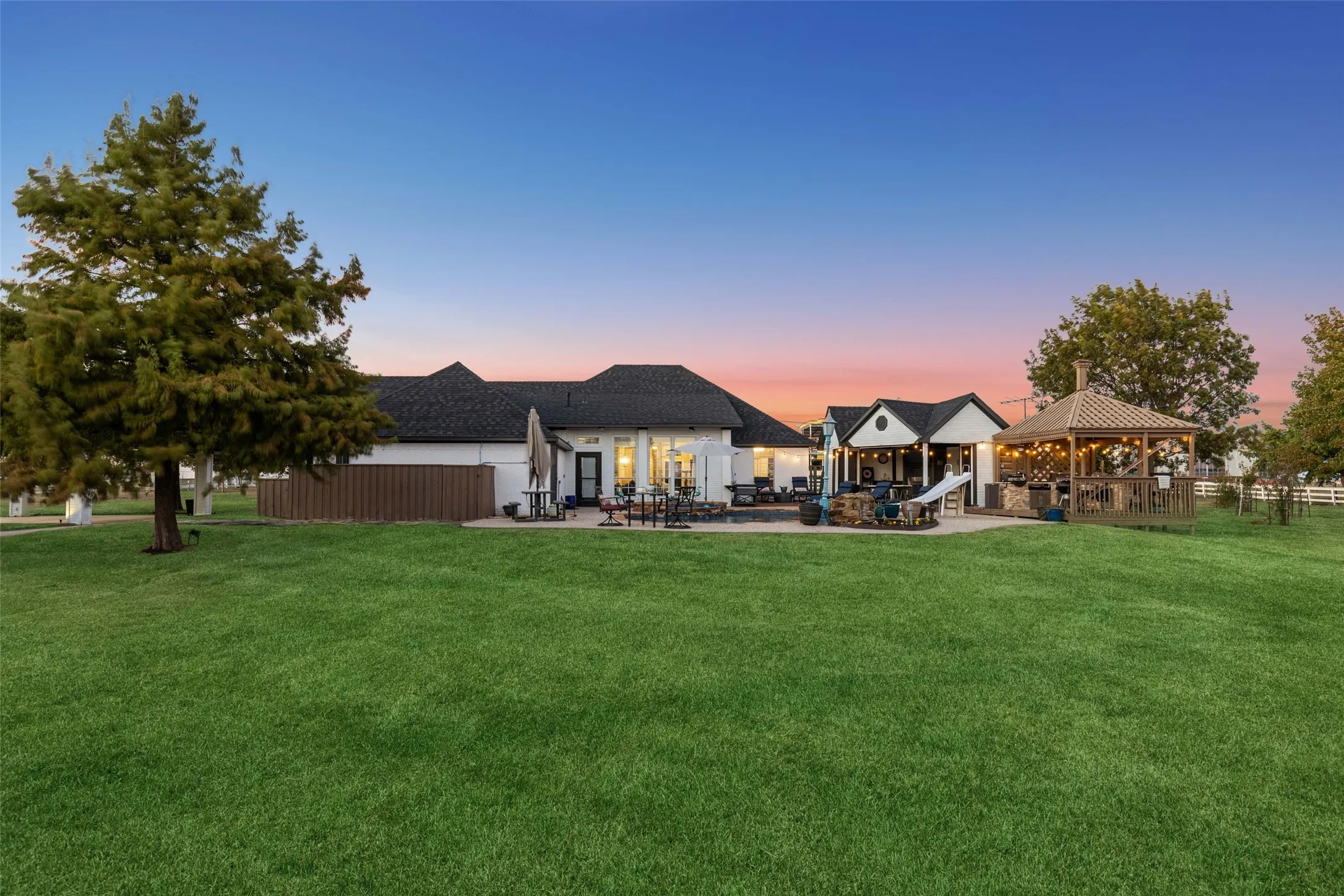 Expansive green lawn with a white exterior residence featuring a dark shingle roof, a covered patio area, and an open-air gazebo