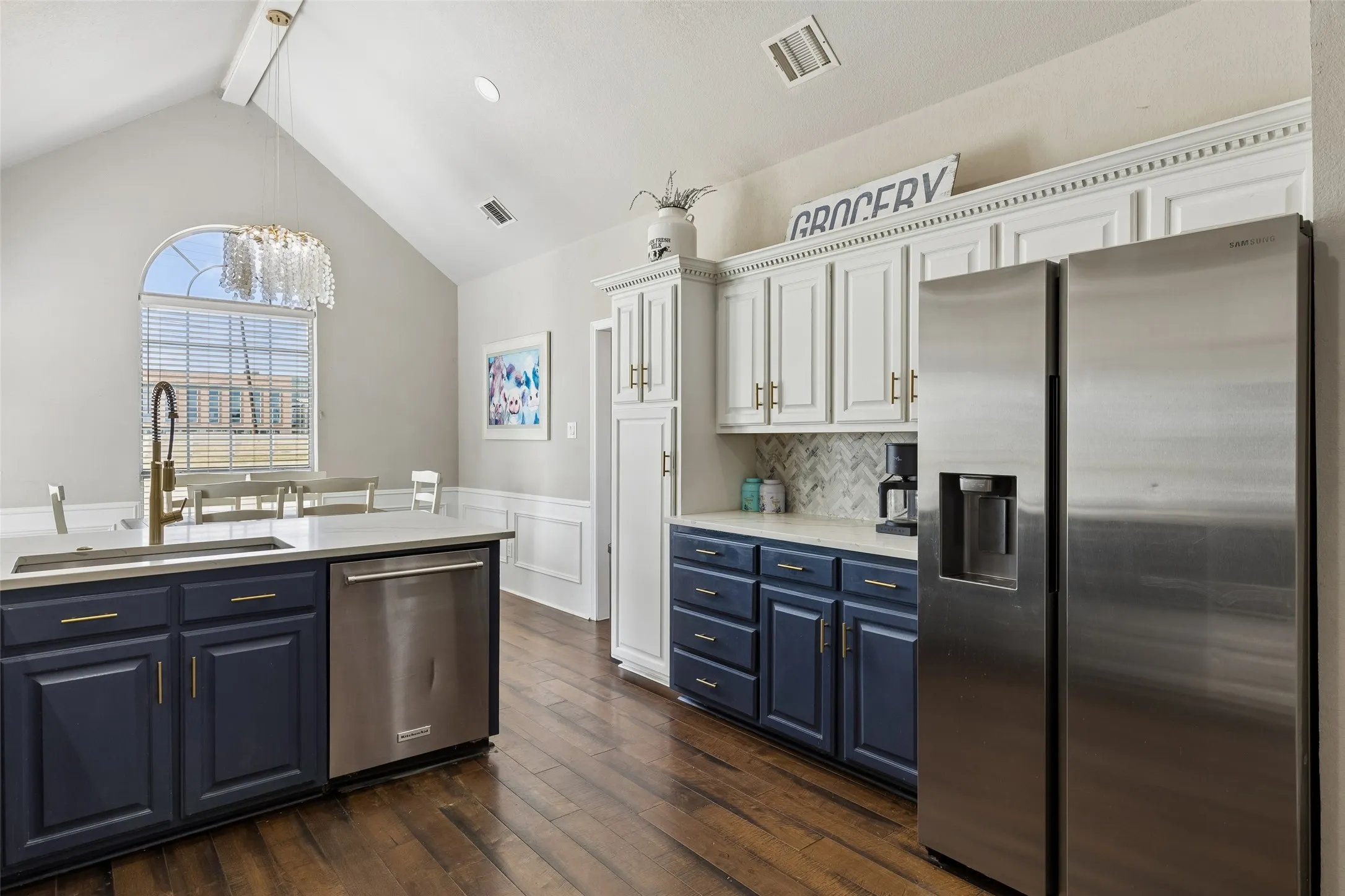 Vaulted ceiling with an arched window, wood-finish flooring, two-tone cabinetry with gold-finish hardware, stainless steel appliances, and a kitchen island with a built-in sink and dishwasher