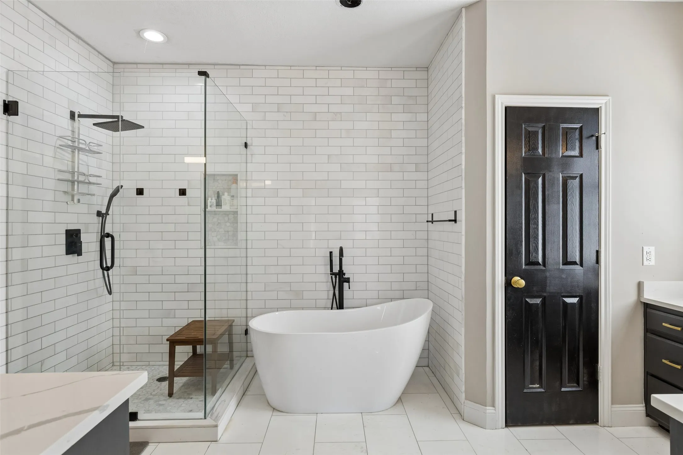 Freestanding soaking tub, glass-enclosed shower with subway tile, built-in shower bench, black hardware, and an adjacent vanity with a white countertop