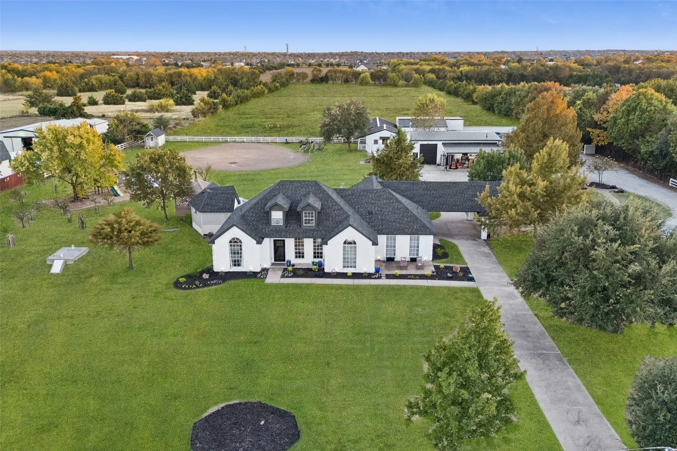 White exterior with dark roof, expansive lawn, concrete driveway, attached carport, and multiple outbuildings