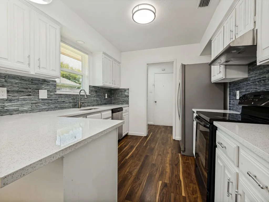 Kitchen featuring white shaker-style cabinetry, light speckled countertops, a textured gray tile backsplash, wood-finish flooring, and stainless steel appliances