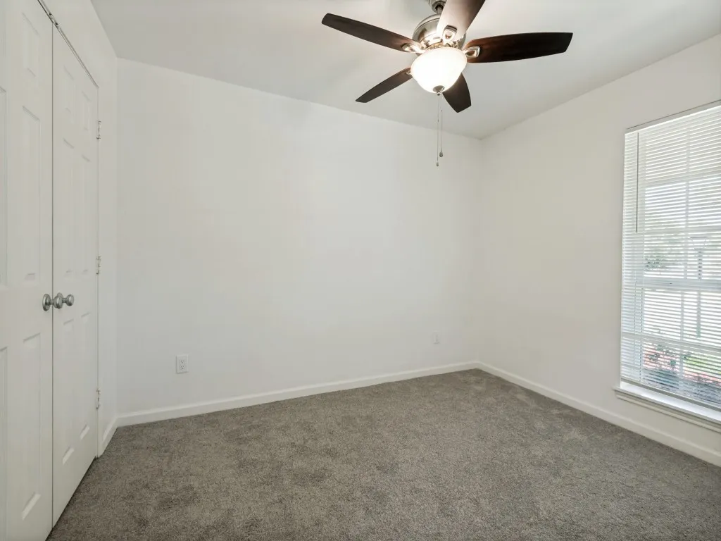 Light gray wall-to-wall carpeting, white painted walls, window with horizontal blinds, bi-fold closet doors, and a ceiling fan with integrated lighting