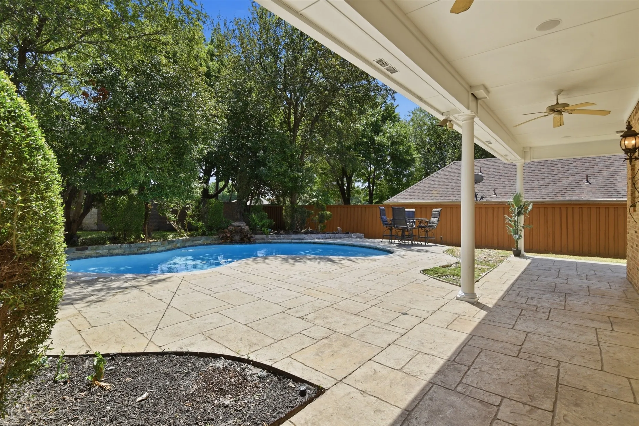 Spacious covered patio with ceiling fans and mounted lighting, expansive stamped concrete patio, inviting swimming pool with waterfall feature, and mature perimeter landscaping