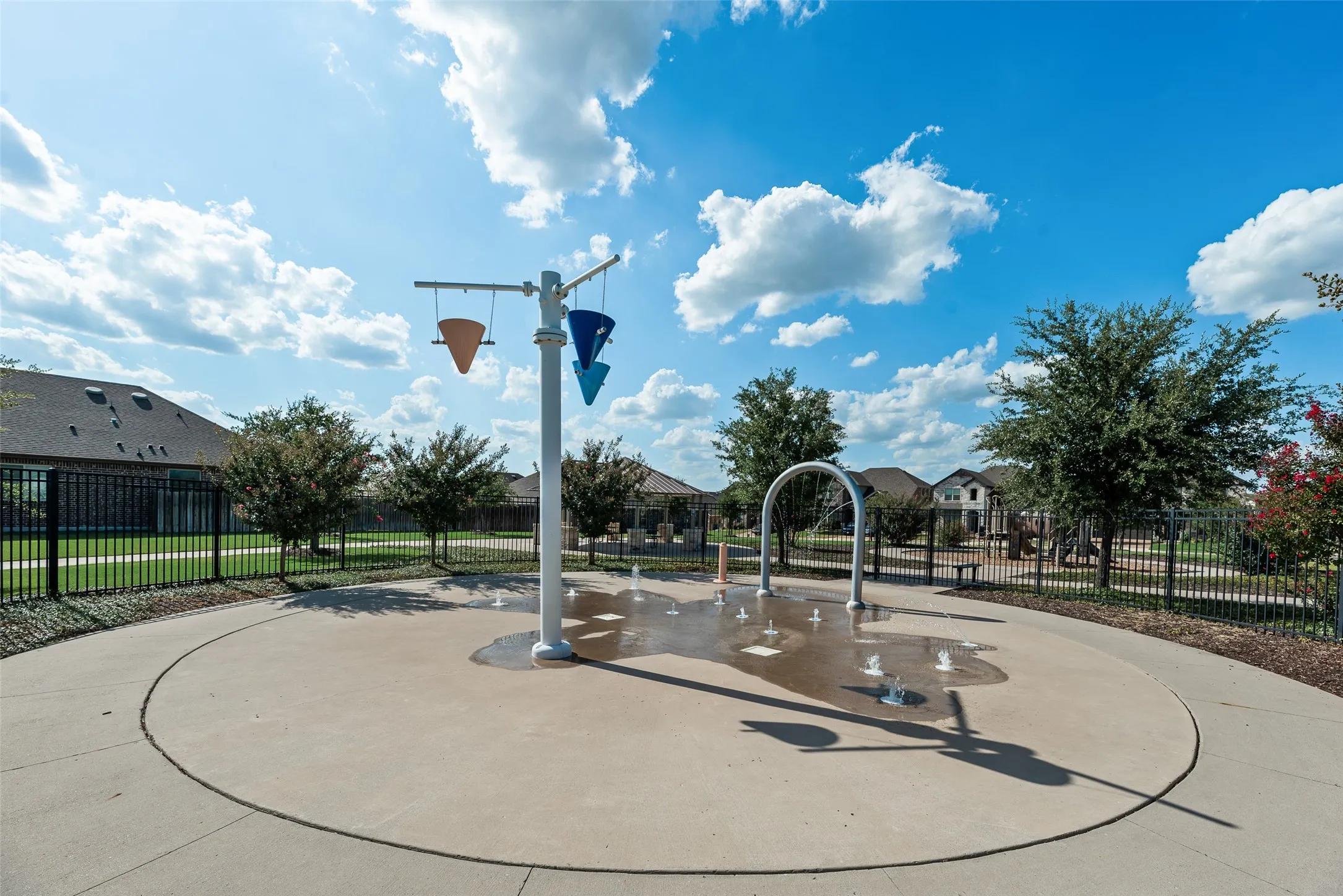 Splash pad with multiple water features, circular concrete surface, and surrounding black metal fencing
