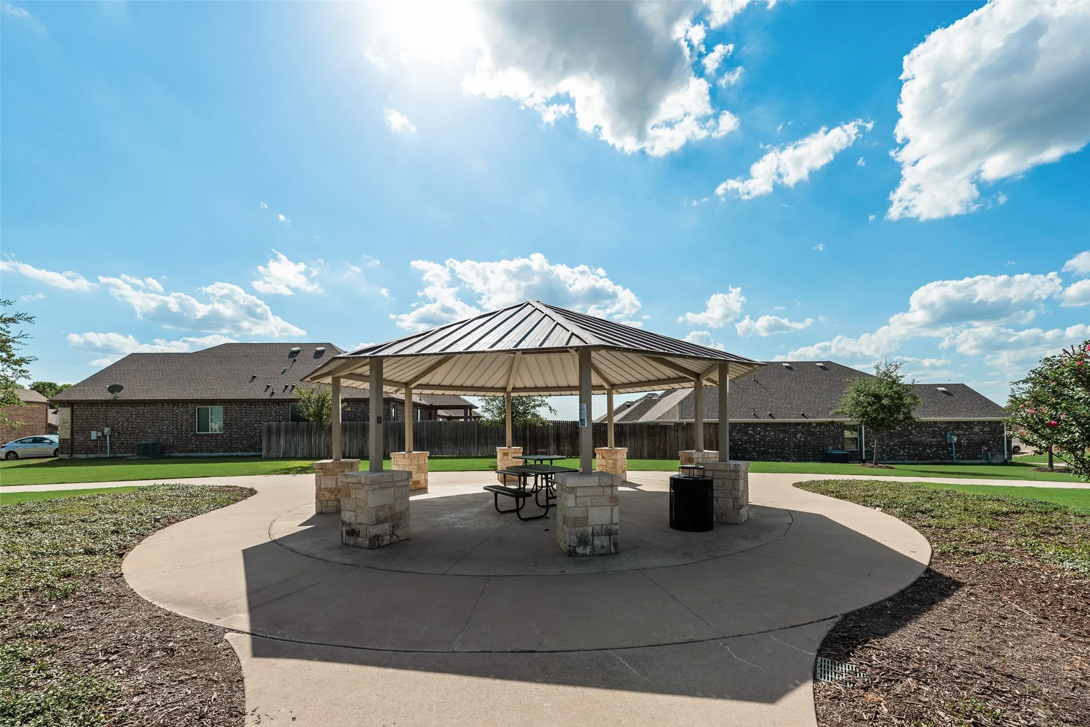 Community gazebo with a metal roof and stone-clad support pillars, surrounded by a concrete pathway and manicured green space