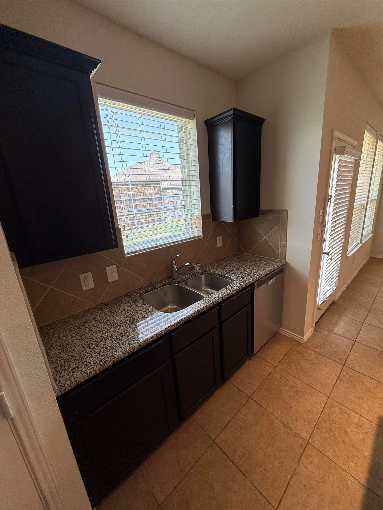 Dark wood-finish cabinetry, granite-style countertops, stainless steel double basin sink, neutral tile backsplash, and ceramic tile flooring